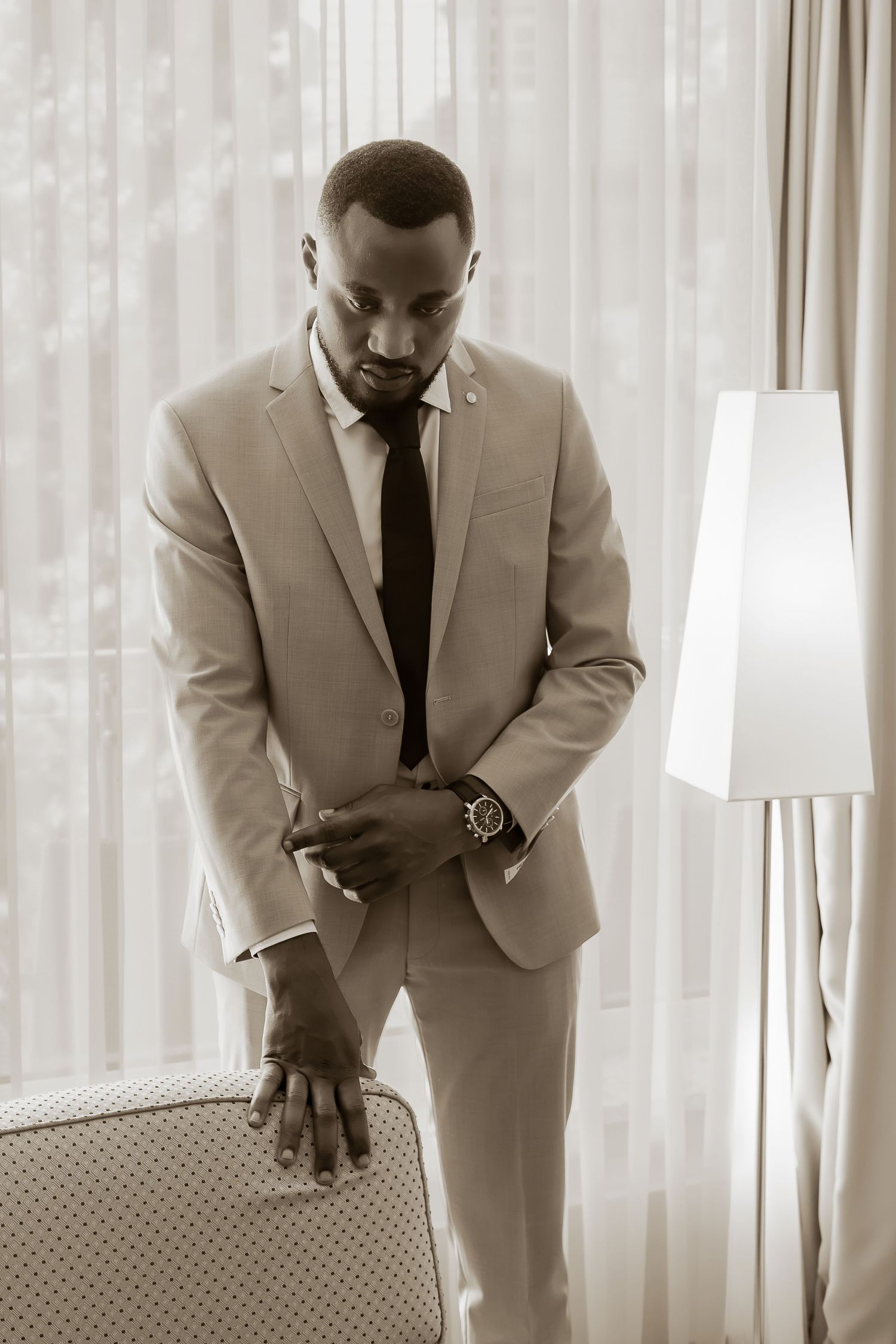 A well-dressed Black man in a beige suit and dark tie adjusts his cufflinks beside a lamp in a bright room.