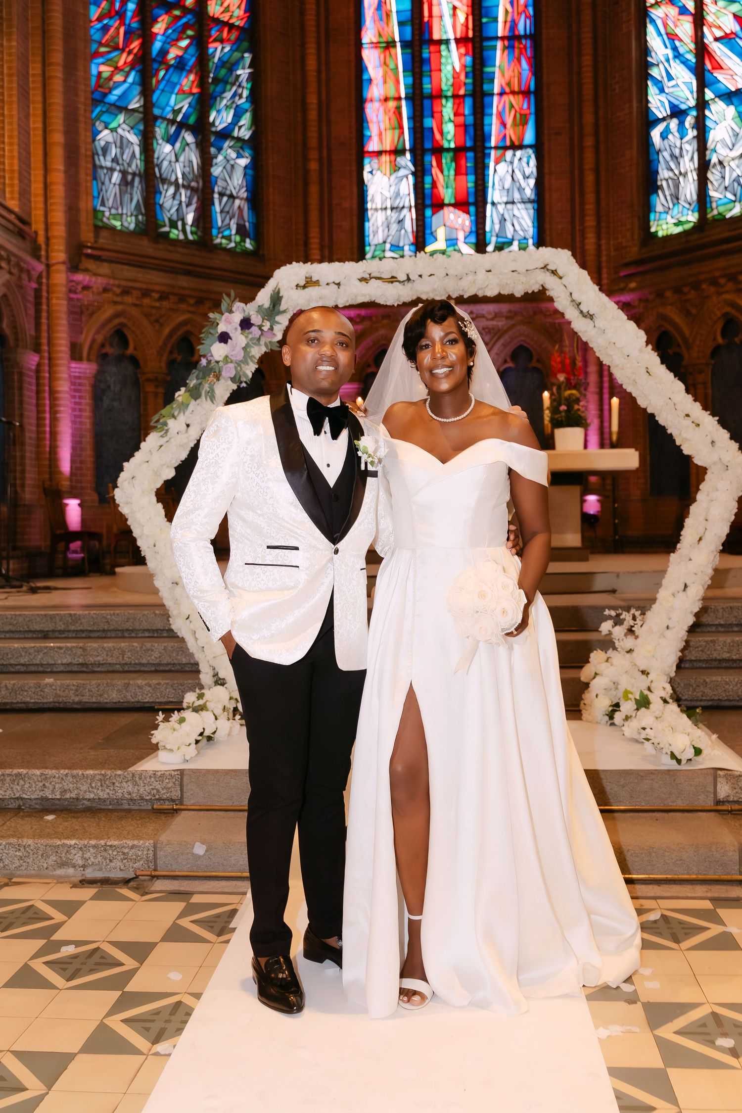 Newlyweds posing under floral arch in stunning church with colorful stained glass windows.