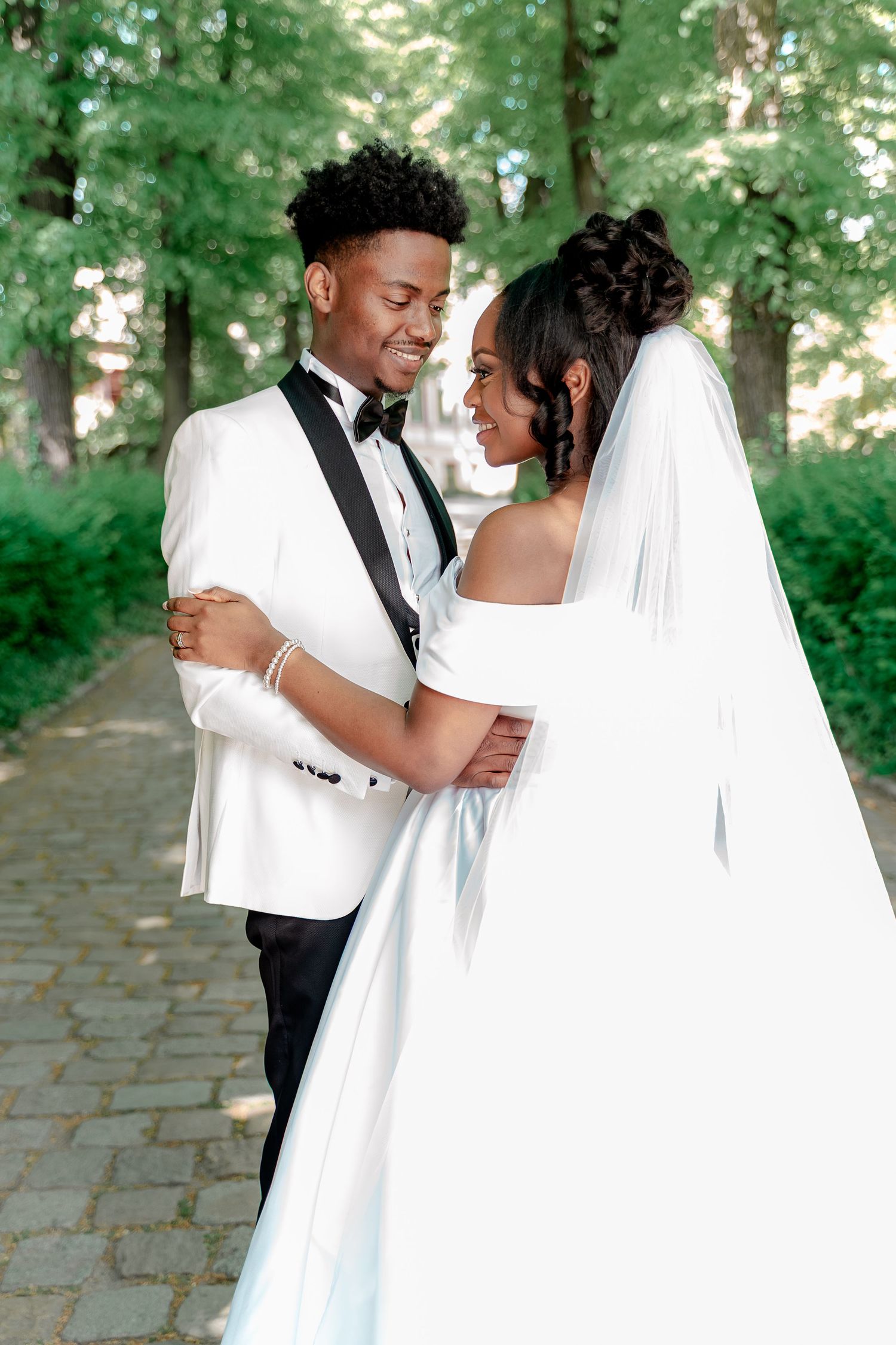A joyful Black couple shares a romantic moment outdoors, groom in white tuxedo, bride in elegant white gown and veil.