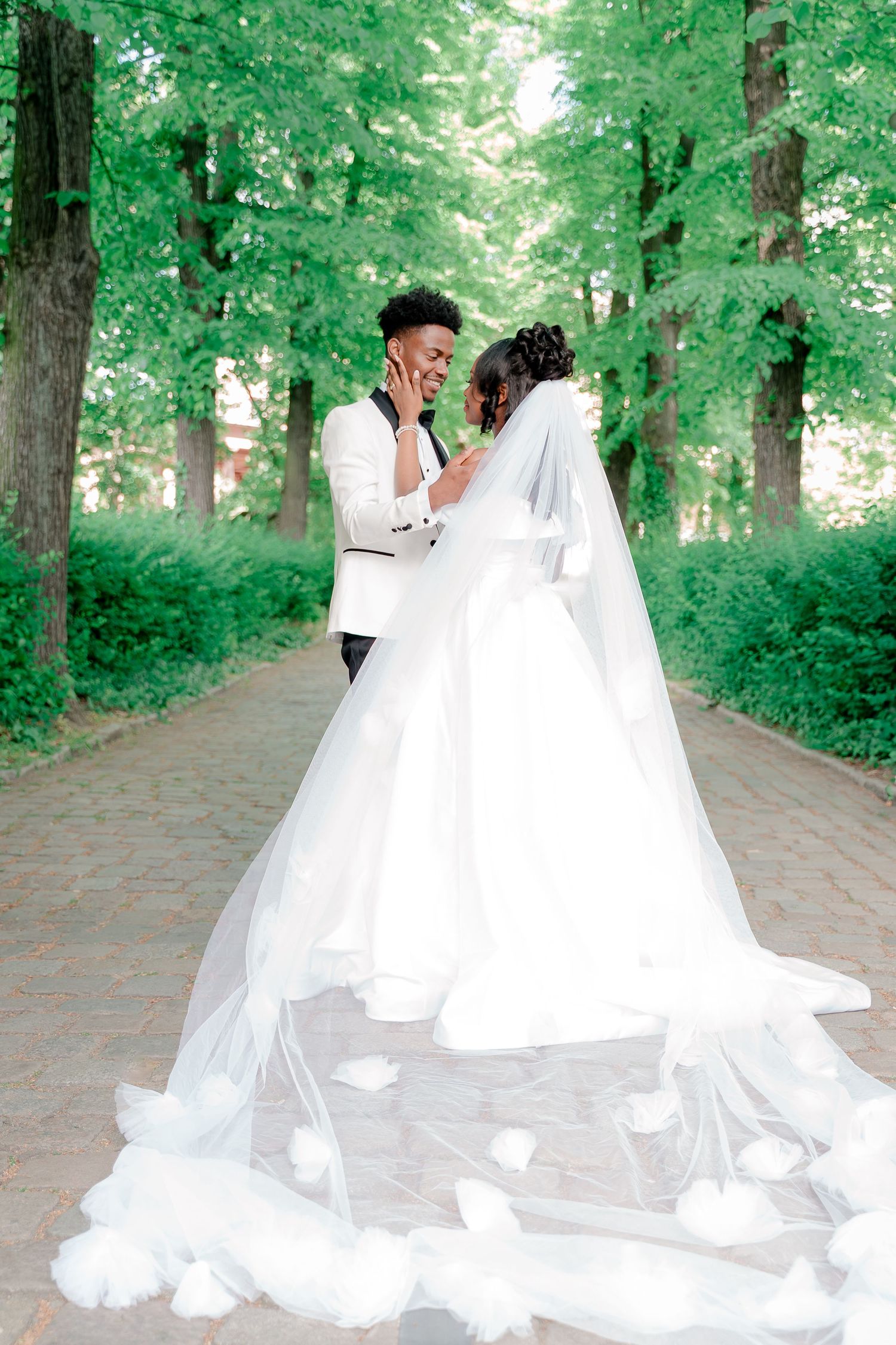 A Black couple shares a romantic moment in a lush green park, bride in a flowing white gown with long veil.