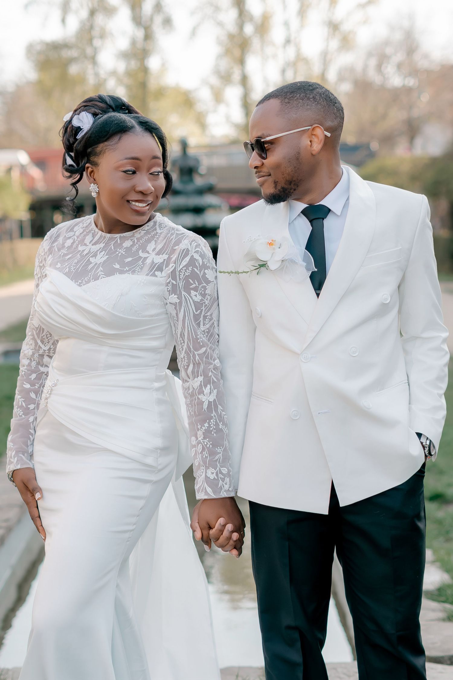 Black bride in long-sleeve lace wedding gown walks hand-in-hand with groom in white jacket outdoors.