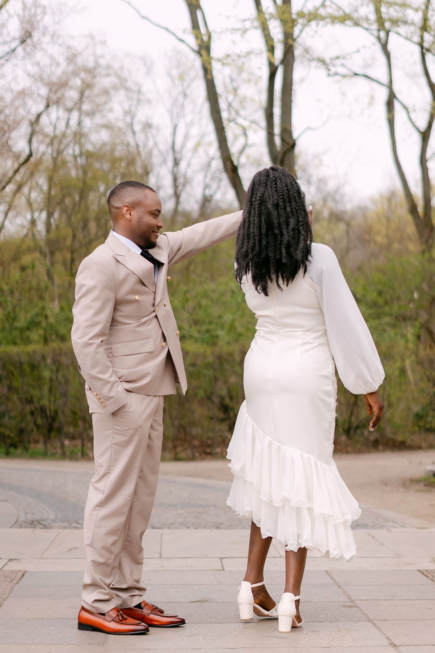 A groom in a beige suit gently touches his brides hair as she stands with her back to him outdoors.