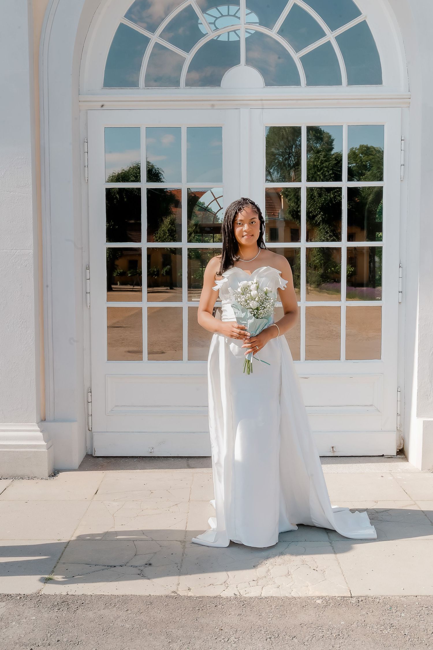 Bride in white strapless gown holding bouquet stands before elegant white French doors on sunny day.