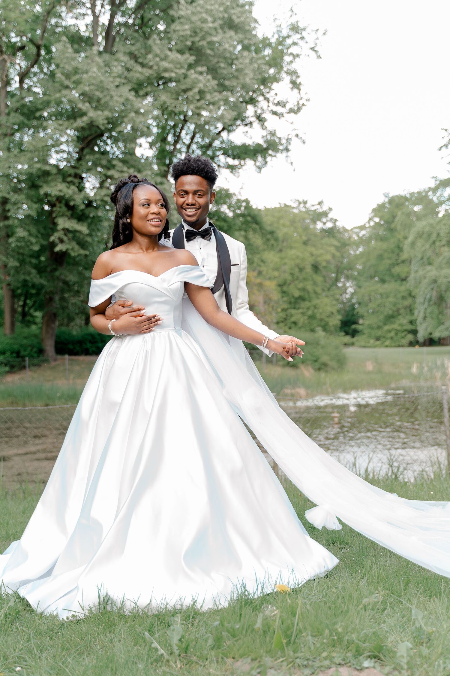 Black groom in white tuxedo embraces bride in off-shoulder satin ball gown near a tranquil pond.
