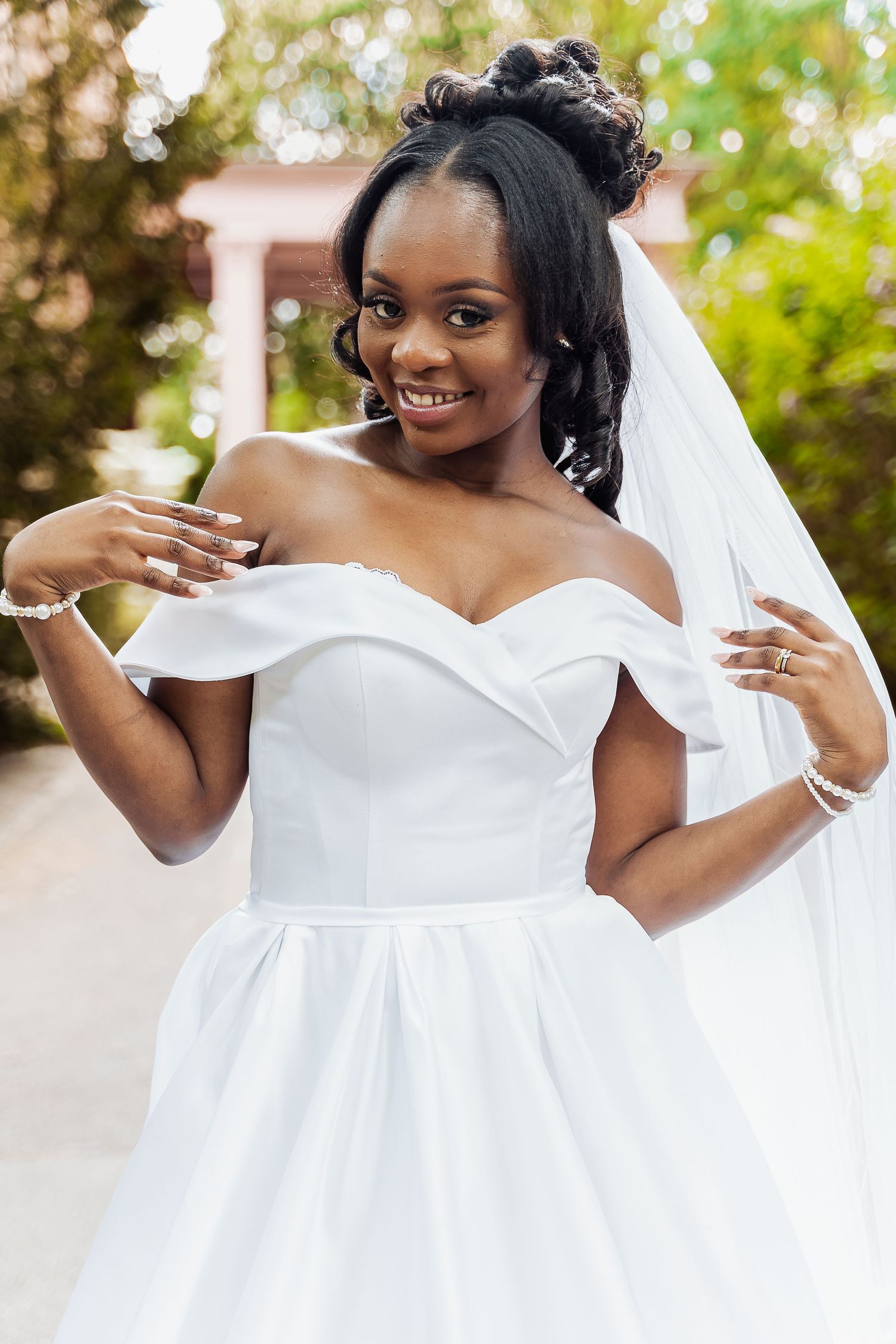 Smiling Black bride in off-shoulder white satin gown and veil poses outdoors in a garden setting.