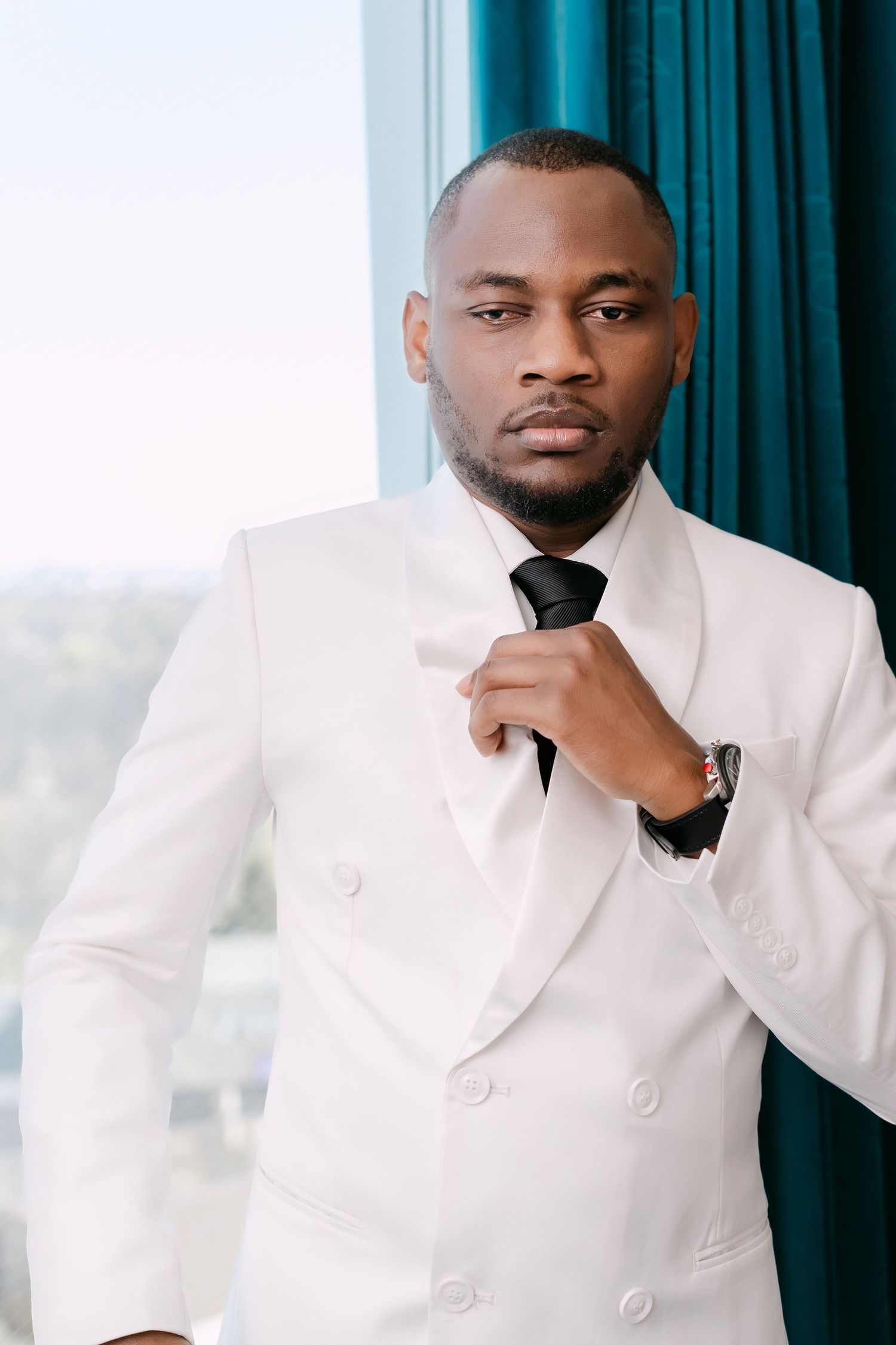 Elegant Black groom in white double-breasted suit adjusting black tie near large window with teal curtains.