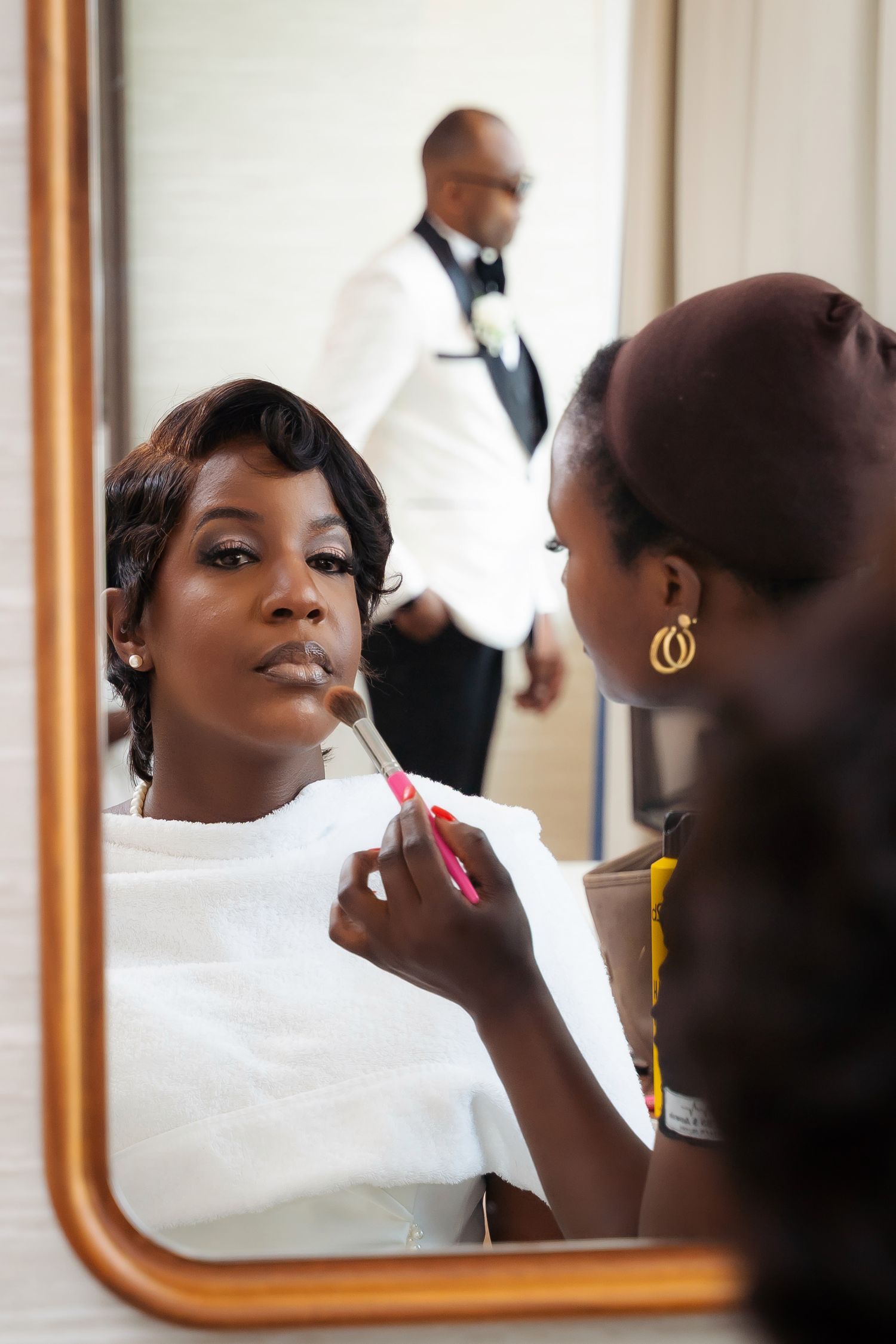 Bride getting makeup applied by artist while reflected in mirror before wedding ceremony.