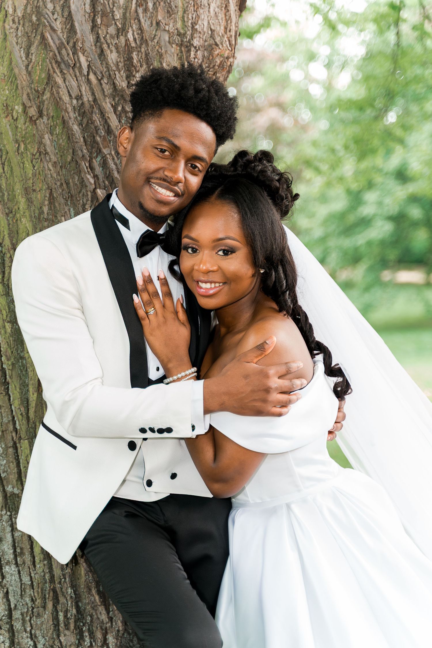 Black bride and groom embrace against a tree, bride in off-shoulder white gown with veil, groom in white tux.
