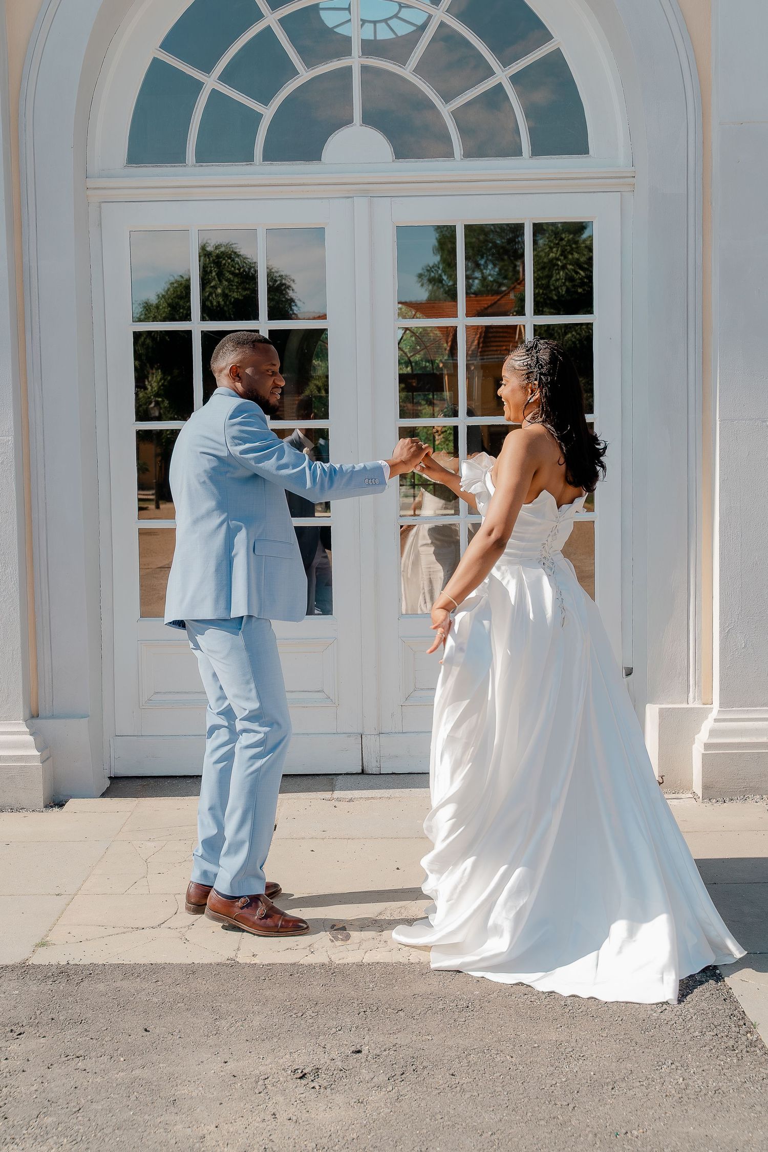 A groom in a light blue suit holds hands with his bride in a white gown, dancing outside an elegant white building.