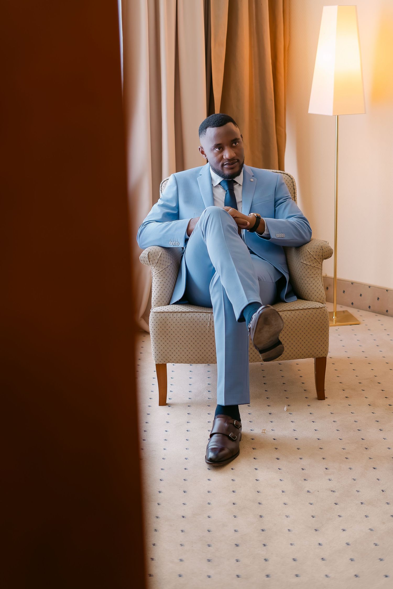 Elegant Black man in light blue suit sitting confidently in armchair, crossing legs, in a warmly lit luxury hotel room.