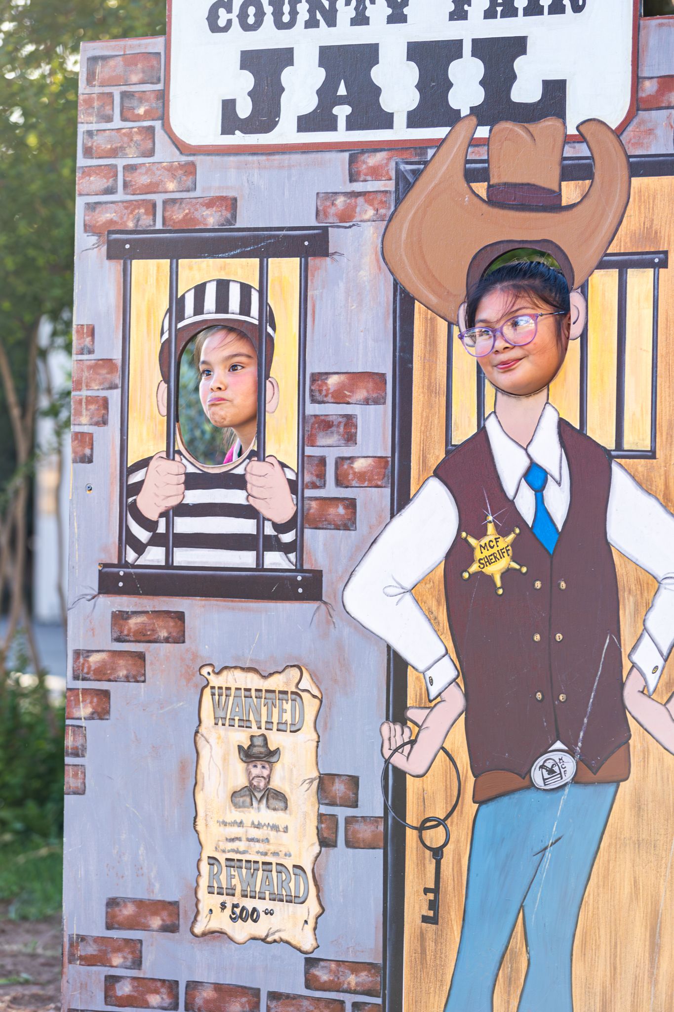 Two children pose at a County Fair Jail photo cutout board, one behind bars and one as sheriff.