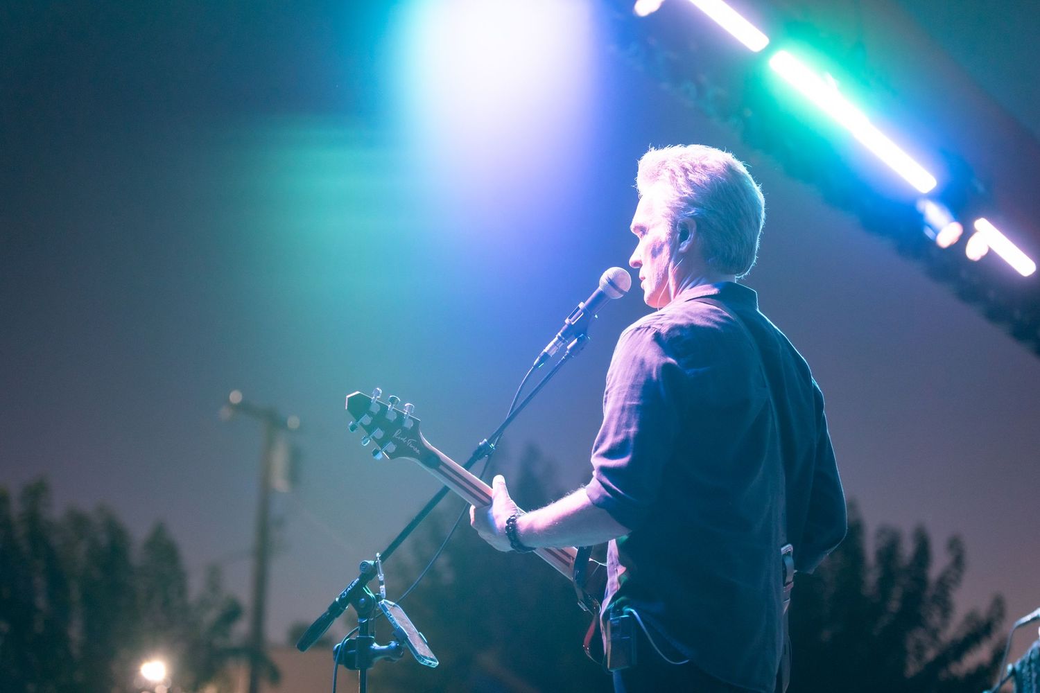 Musician performing on stage with guitar at an outdoor concert under colorful blue and green stage lights.