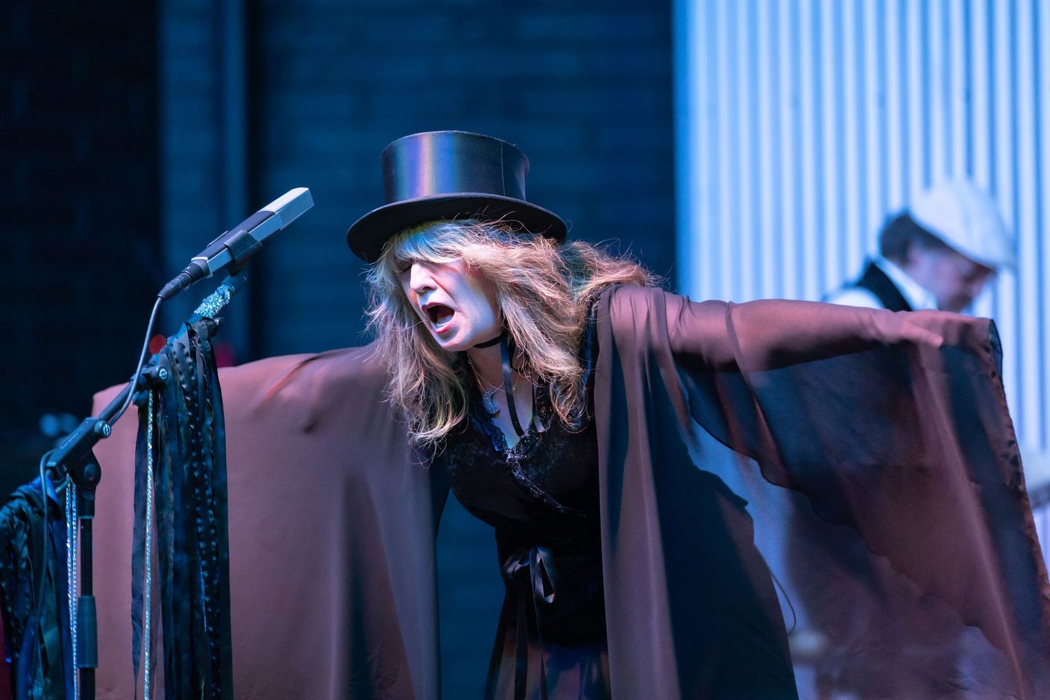 Female performer in a black top hat and cape sings passionately on stage under dramatic blue lighting.