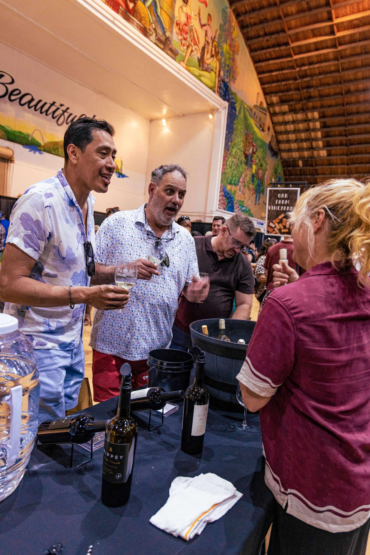 People enjoying a wine tasting event at a vineyard booth with bottles and spittoons on the table.