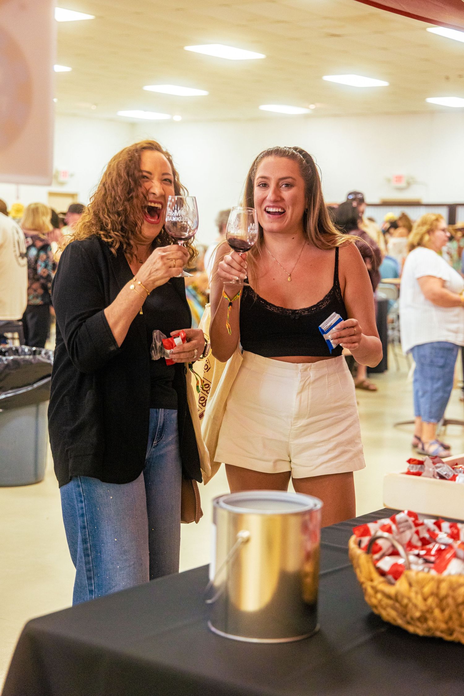 Two women laughing and holding red wine glasses at a lively indoor wine tasting festival event.