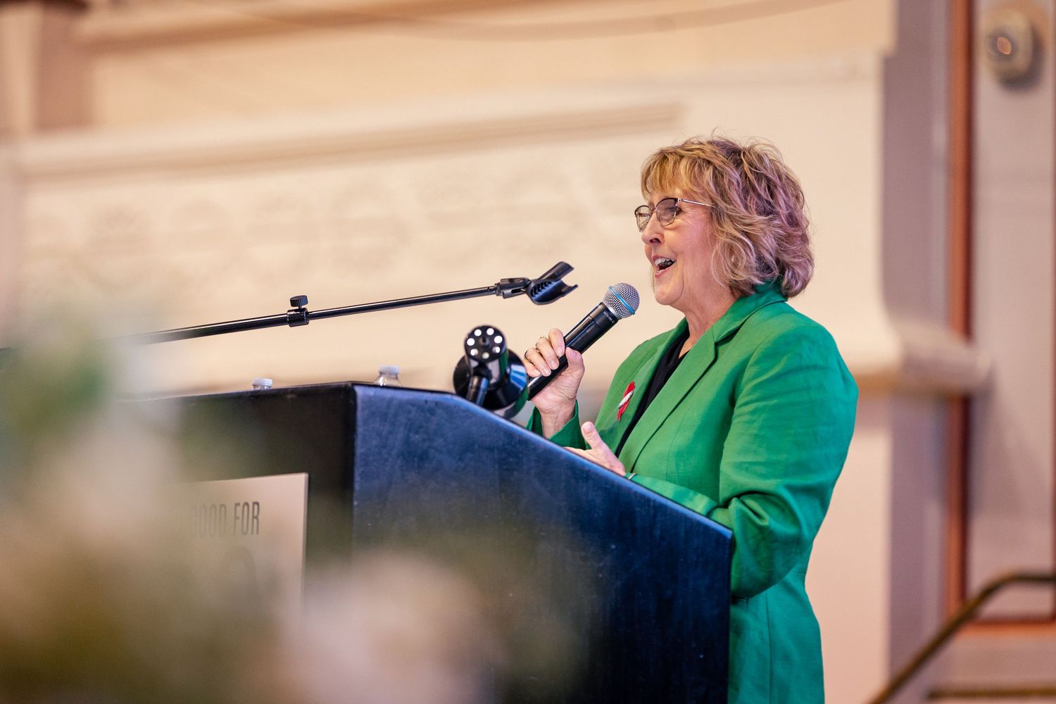 Woman in green blazer speaking at podium with microphone during formal event or conference.
