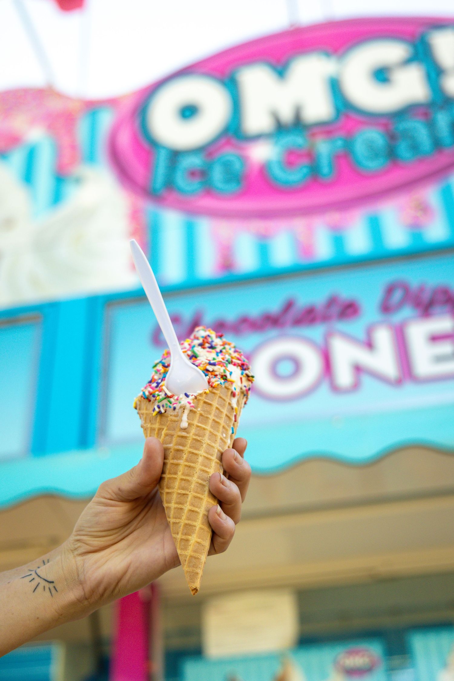 Hand holding a sprinkle-topped ice cream cone in front of a colorful OMG Ice Cream shop.
