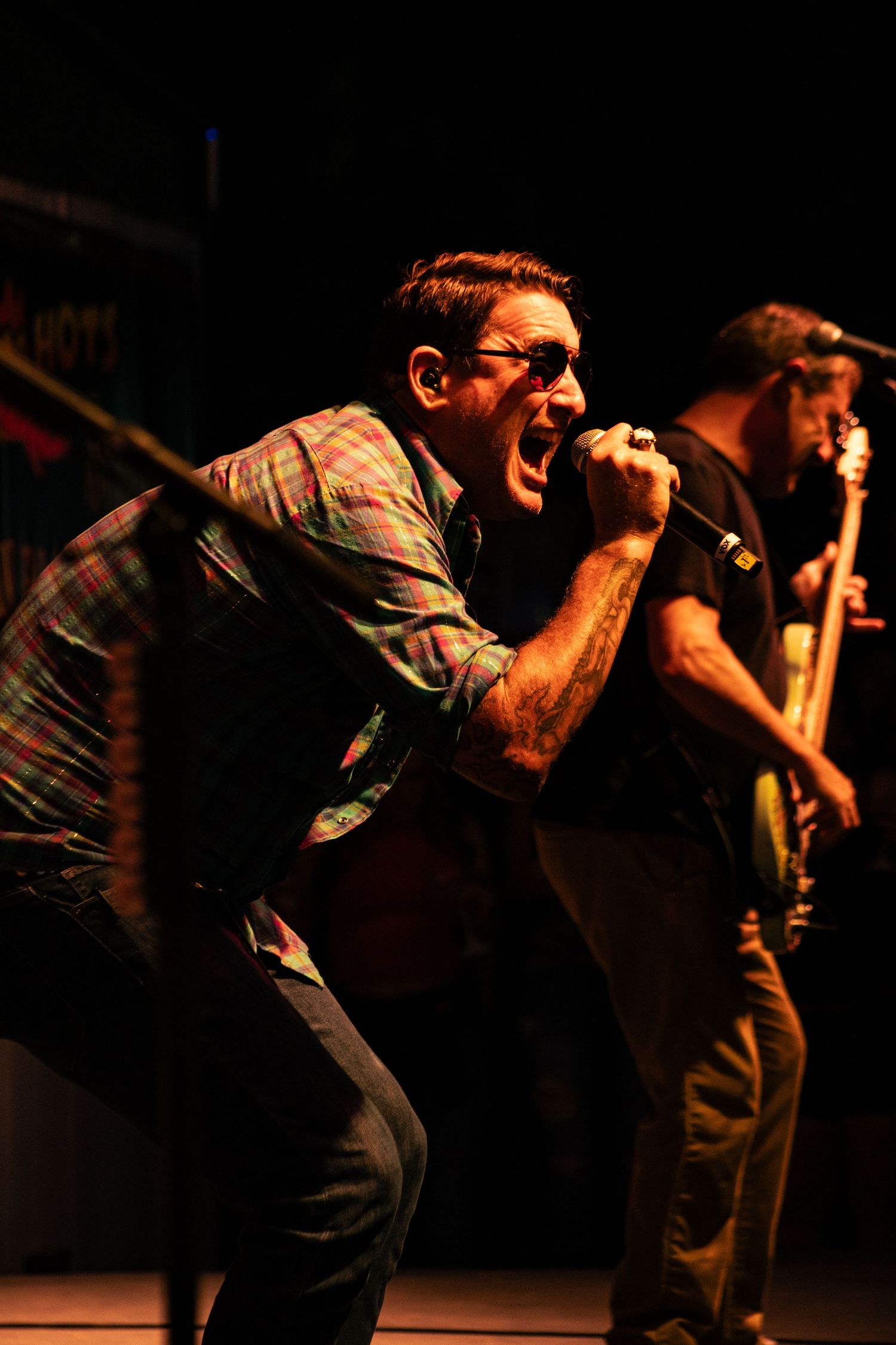 A passionate male vocalist screams into a microphone on stage, wearing a plaid shirt, with a guitarist visible behind him.