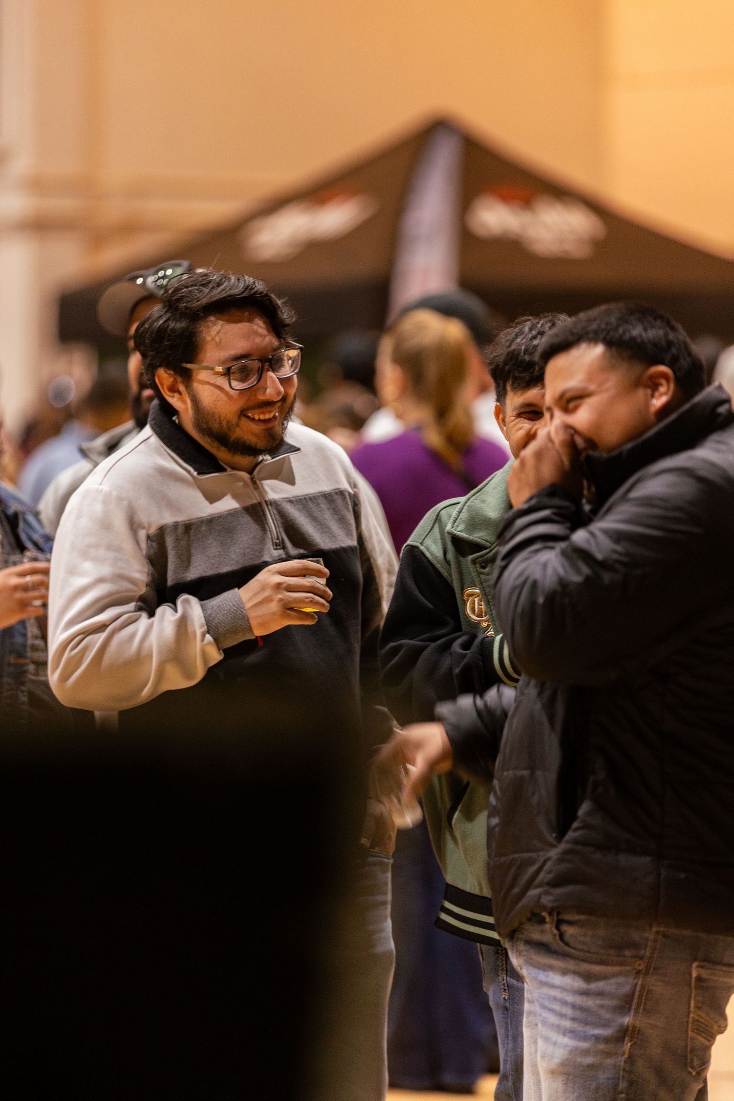 Two men laughing and socializing at an indoor event with a dark tent in background.