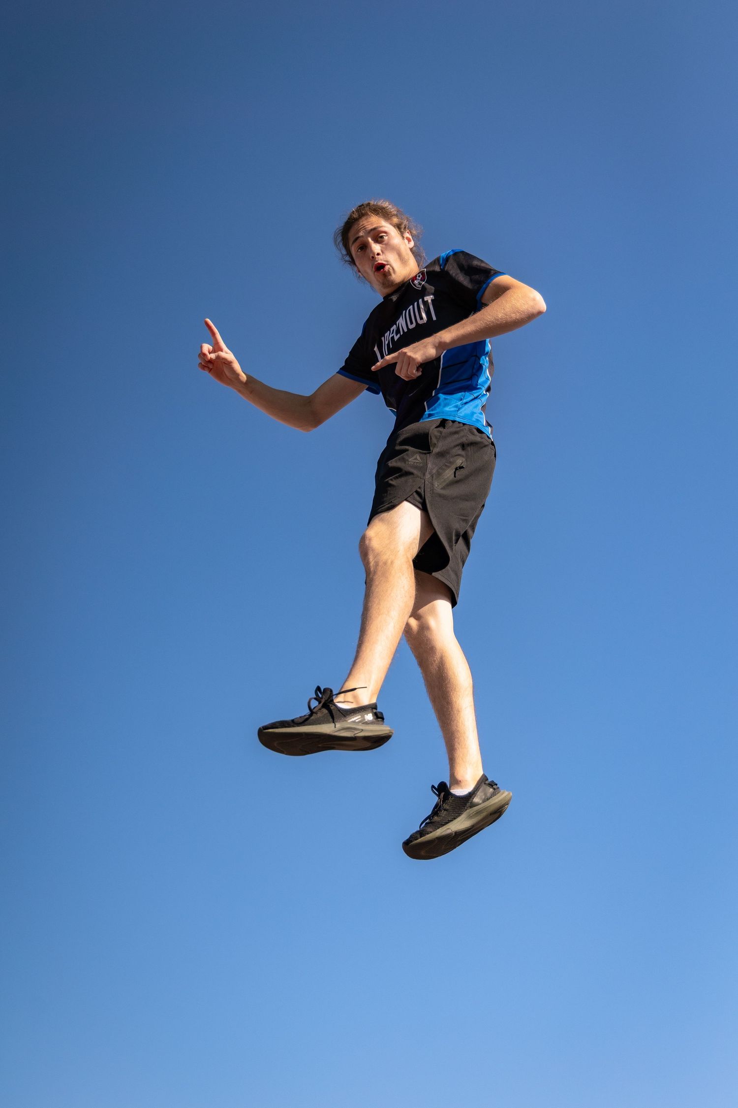 A teenage boy jumps high against a clear blue sky, striking a dynamic mid-air pose outdoors.