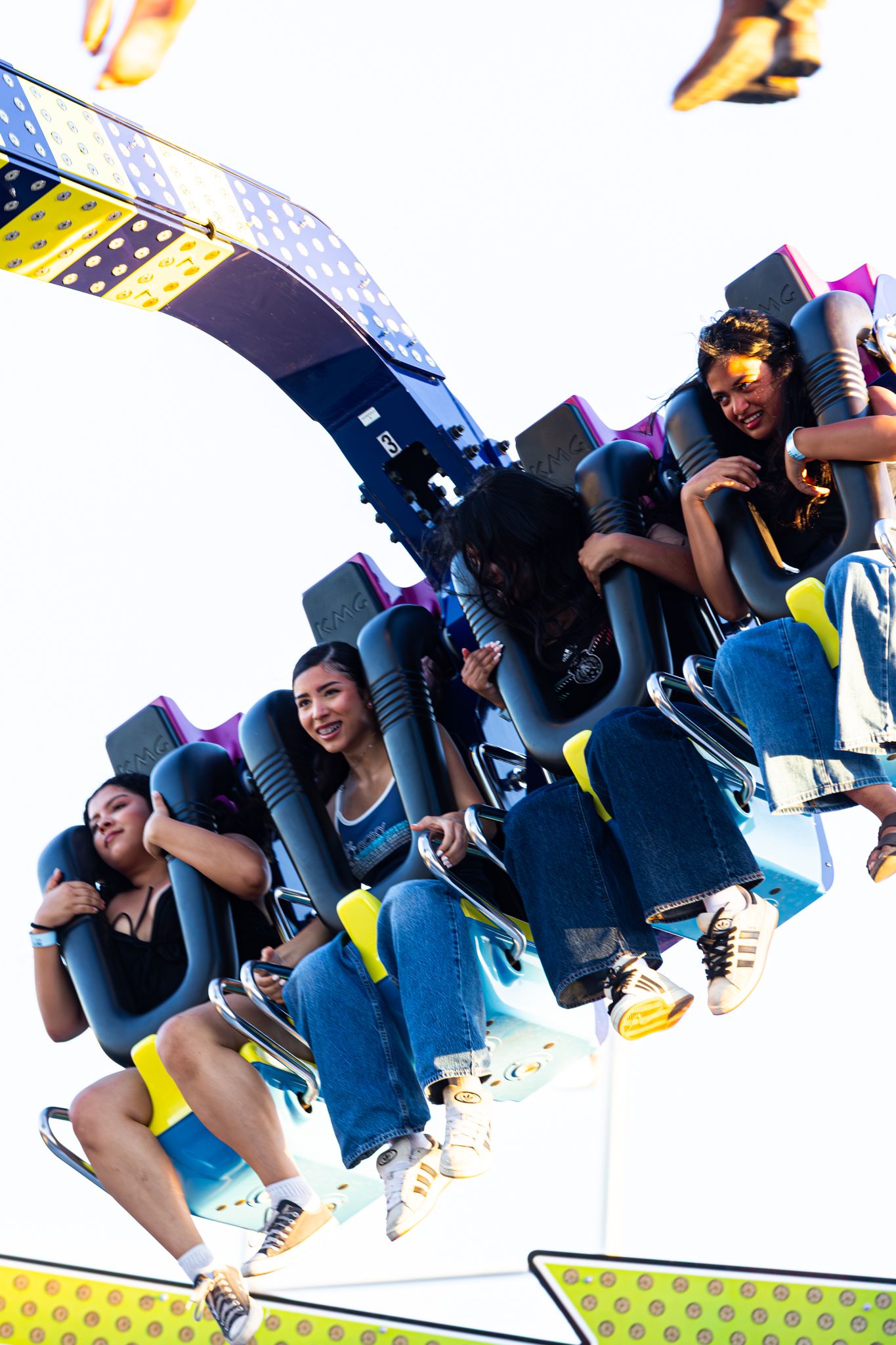 Excited riders on a spinning amusement park thrill ride, laughing and smiling against a bright sky at a fair.