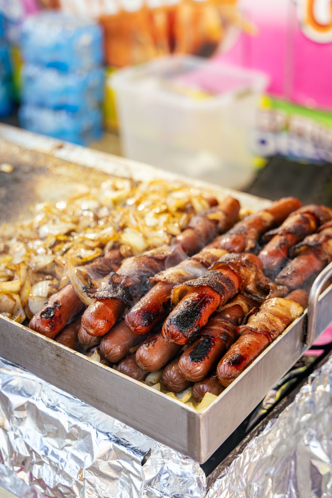 Bacon-wrapped sausages and caramelized onions grilling on a foil-lined tray at a street food market.