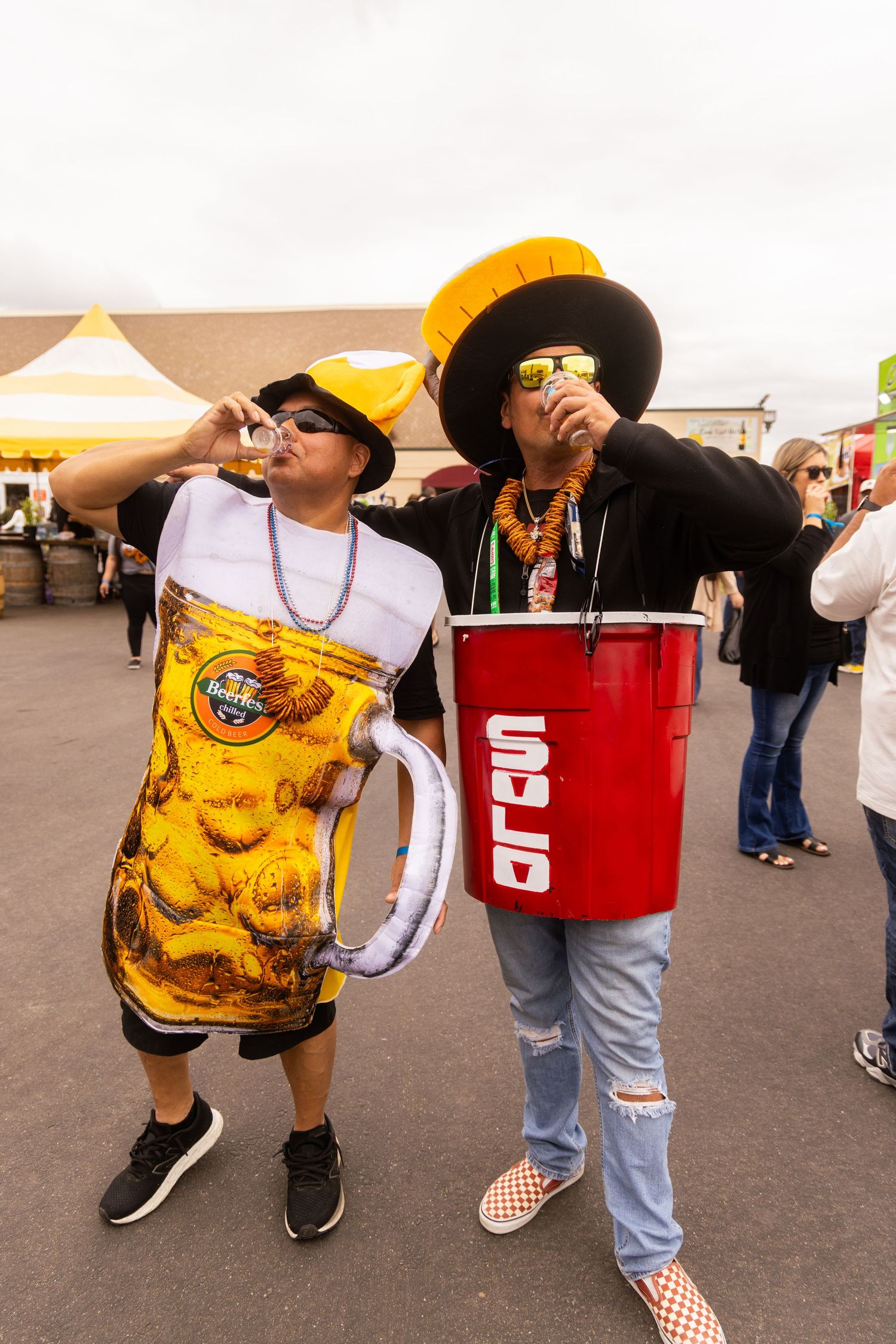 Two people in fun beer-themed costumes drinking at an outdoor Oktoberfest celebration event.
