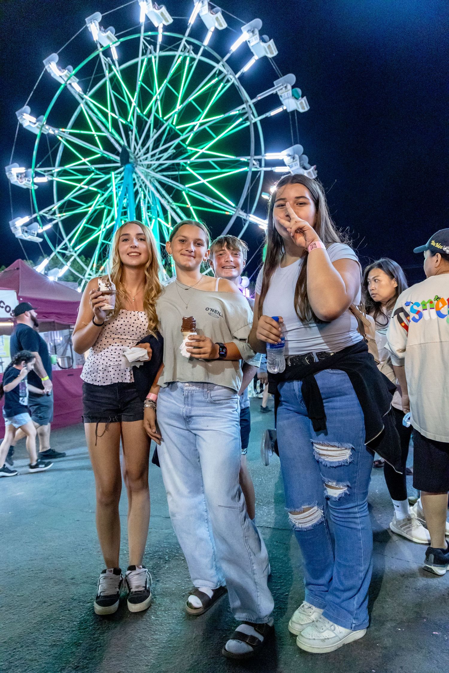 Group of young women smile and pose in front of a glowing neon Ferris wheel at a night carnival fair.