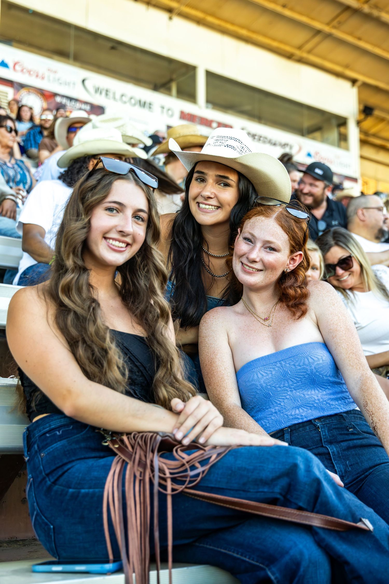 Three smiling women wearing cowboy hats and jeans sitting together in stadium bleachers at a rodeo event.