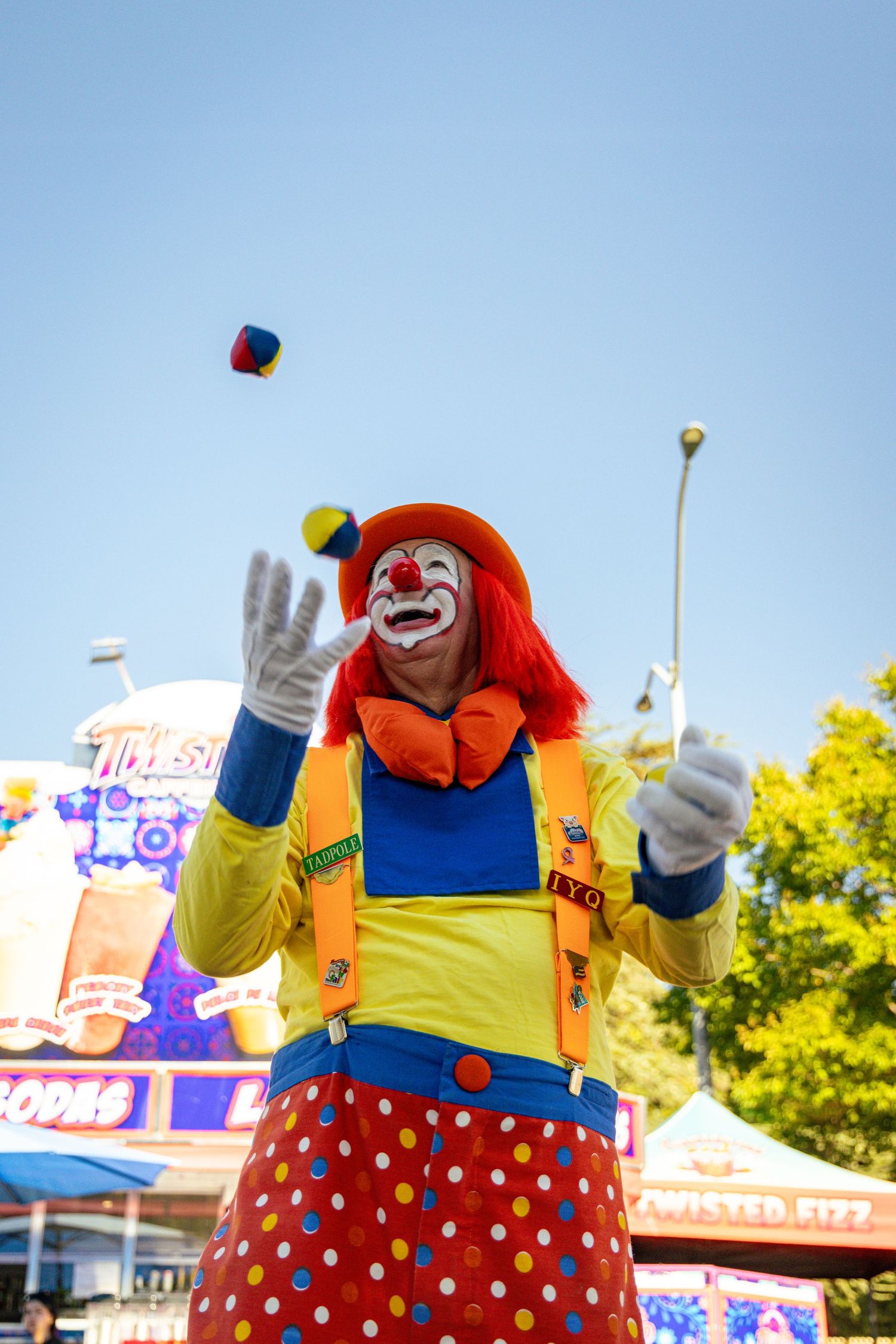 Colorful clown in red wig juggling balls at a sunny outdoor carnival fair.