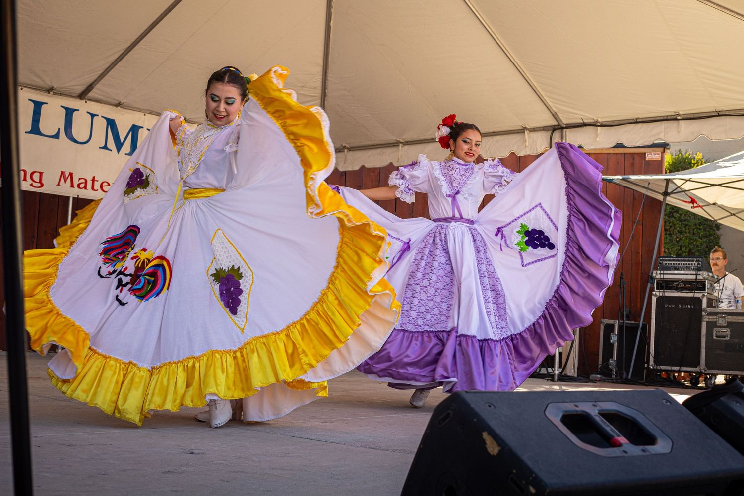 Two women perform traditional Mexican folkloric dance in colorful white and purple dresses on an outdoor stage.