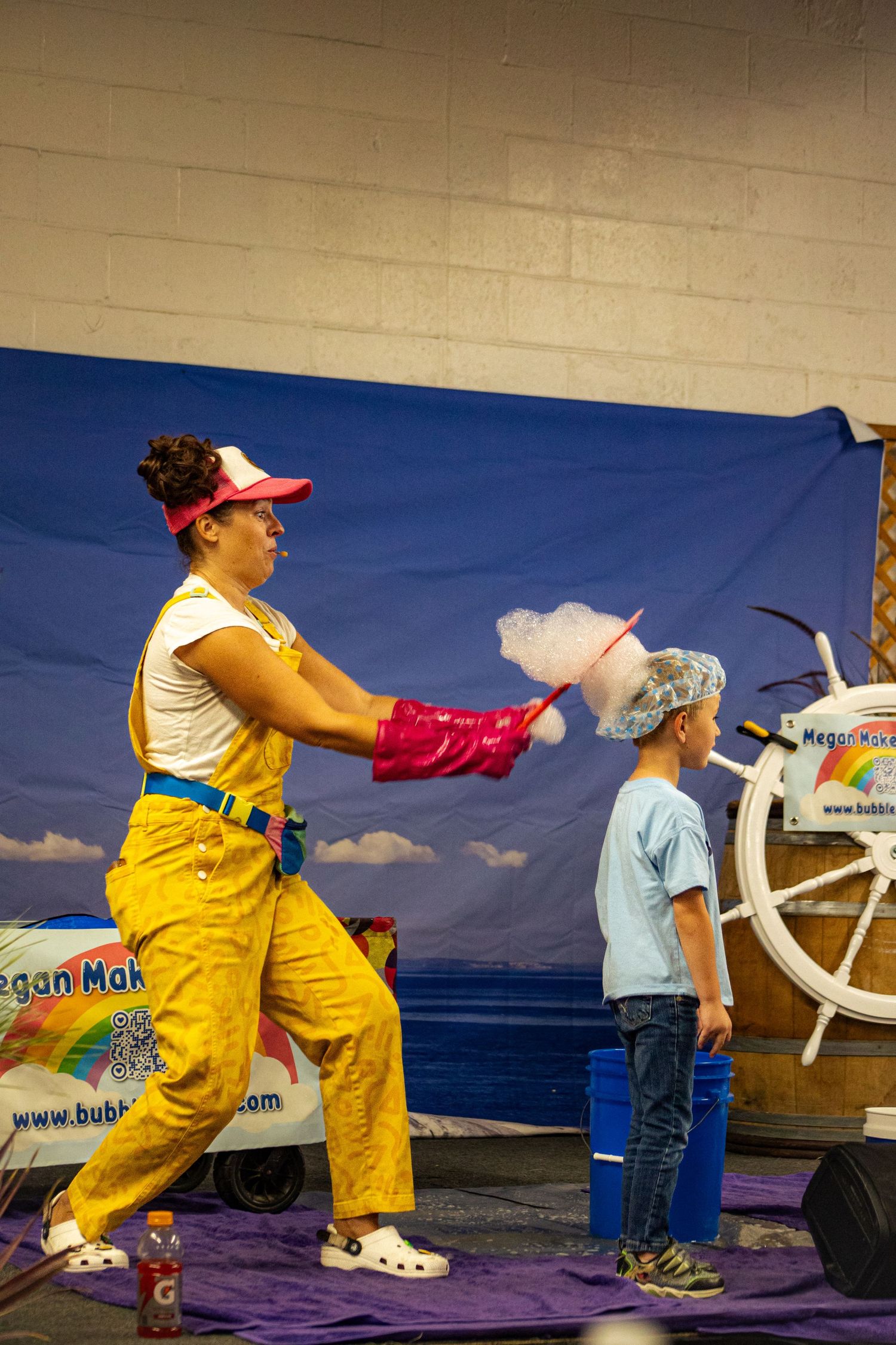 A clown in yellow overalls makes cotton candy for a child at an indoor fair or carnival event.