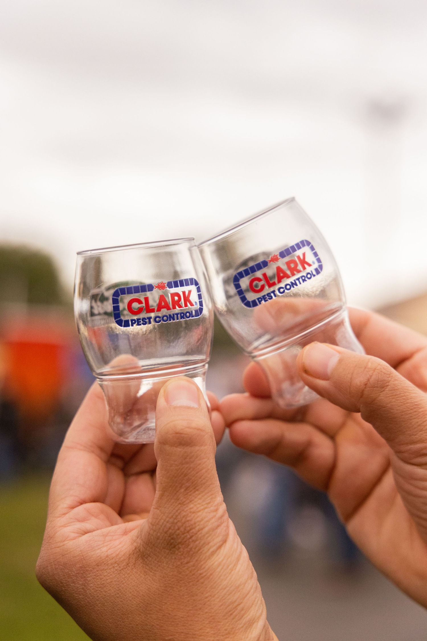 Two hands clinking small Clark Pest Control branded tasting glasses at an outdoor event.