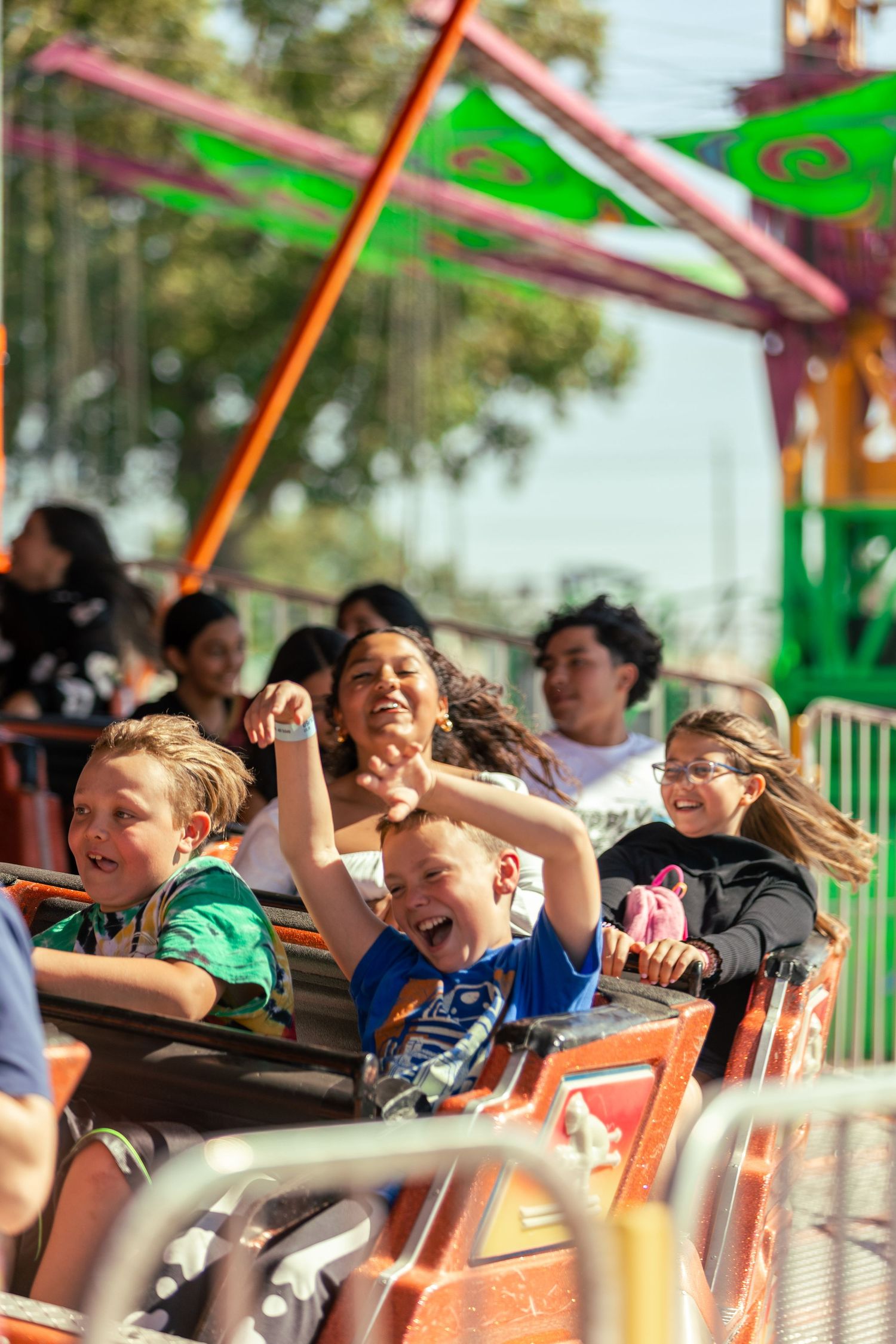 Excited children laugh and cheer with hands raised while riding a colorful carnival roller coaster ride.