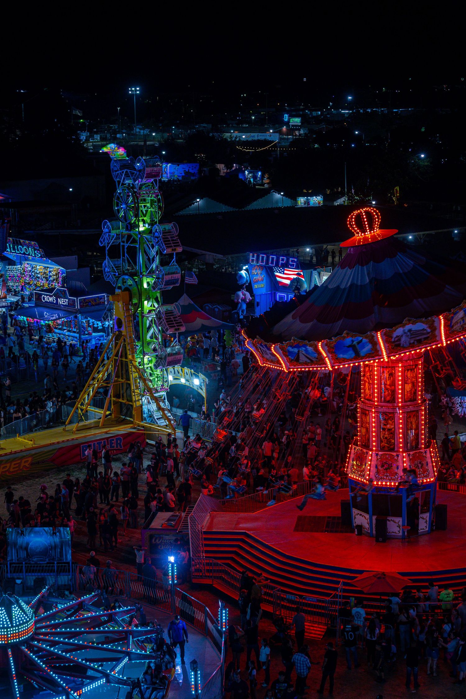Aerial night view of a colorful carnival midway with glowing rides and crowds.