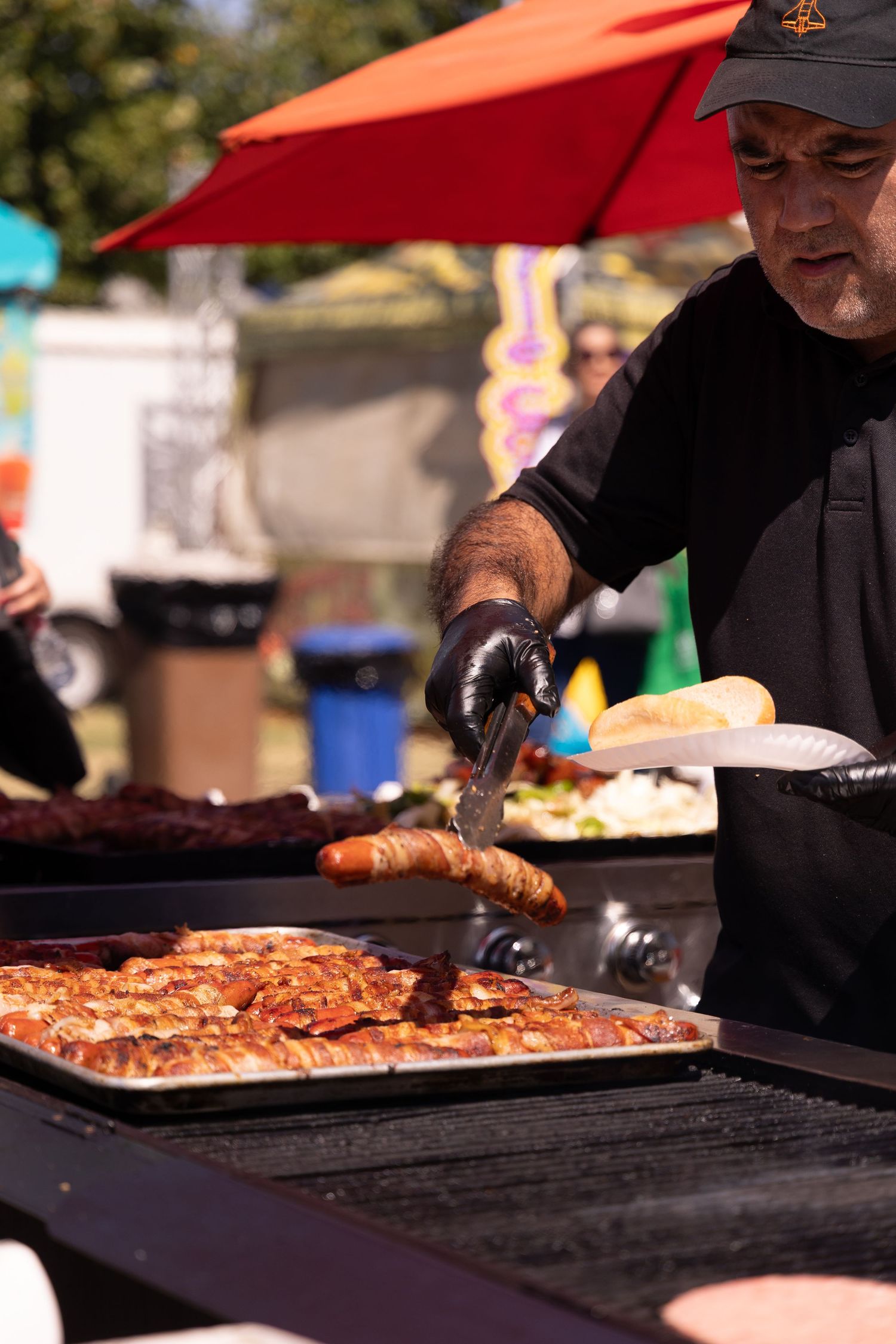 Vendor in black grilling sausages and bacon at an outdoor fair food stand.