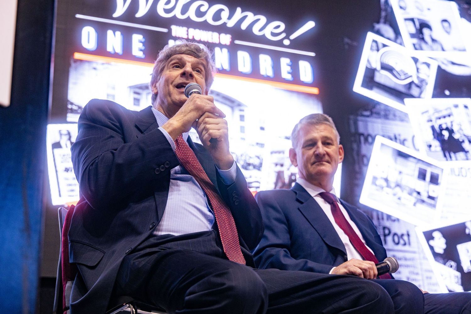 Two men in suits sit on stage at a Welcome event, one speaking into a microphone under colorful event signage.