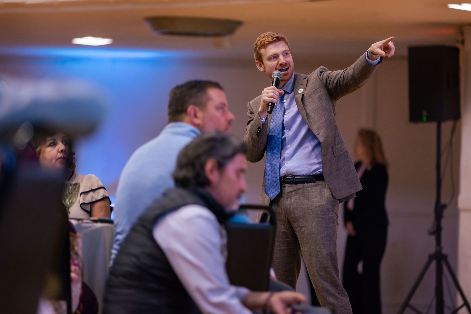 A man in a blazer speaks into a microphone while pointing at the audience during an indoor event.