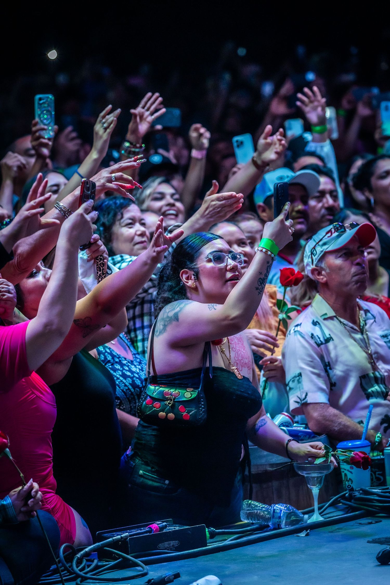 Excited concert crowd raises hands and phones under colorful lights as a woman holds a rose up front.