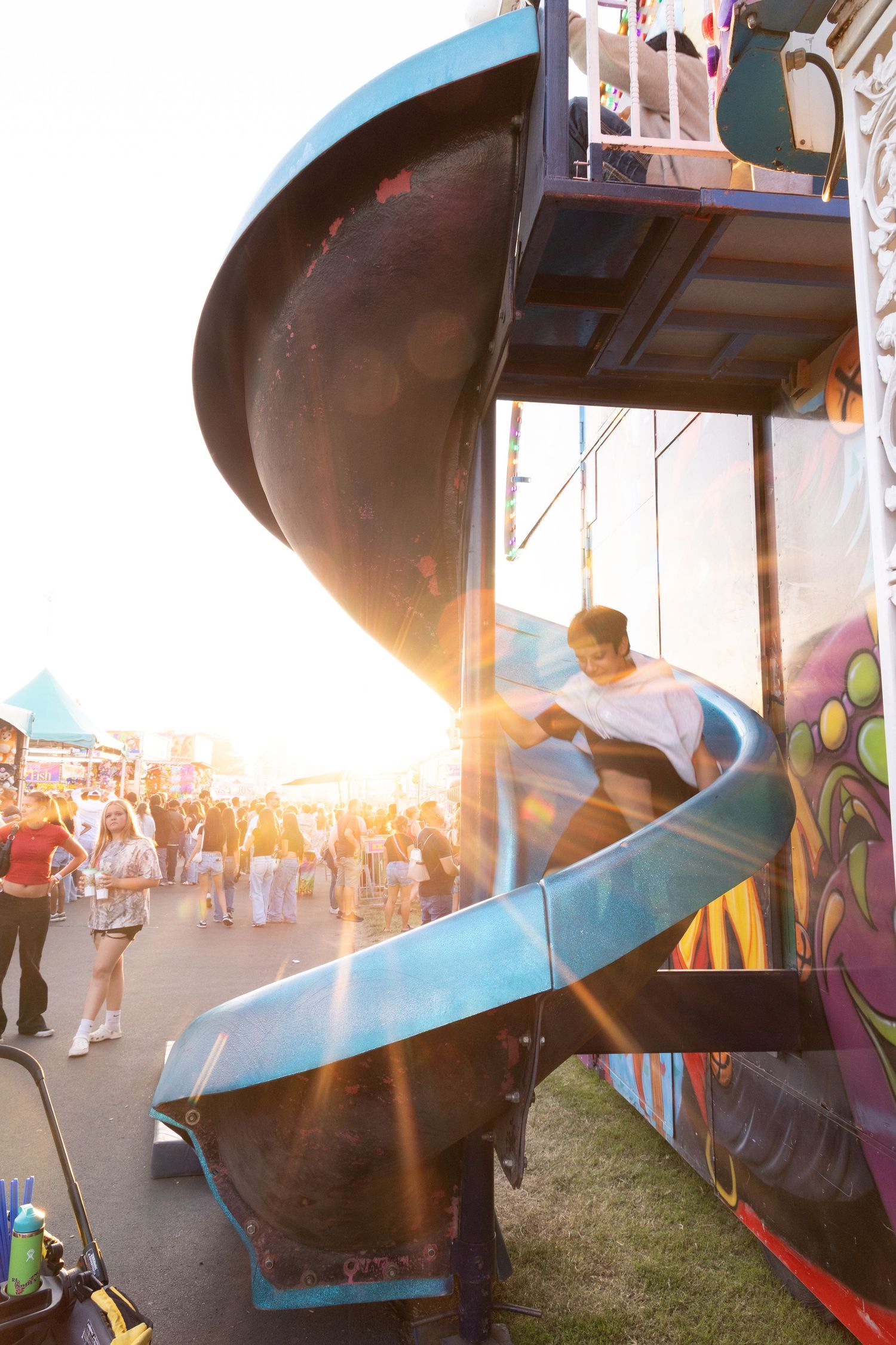 Person sliding down a spiral blue slide at a sunny outdoor carnival with lens flare.