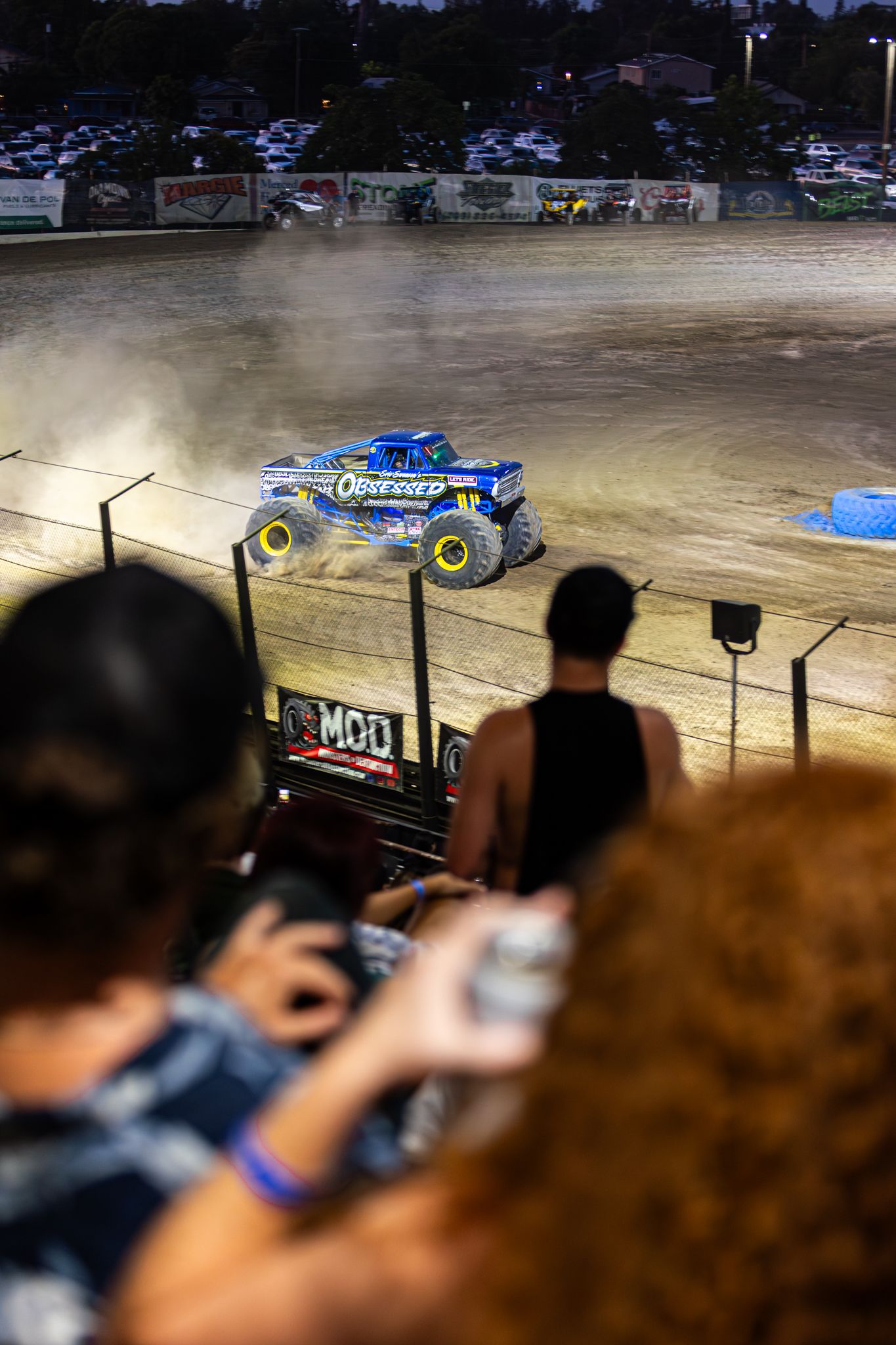 Monster truck Obsessed races through dirt arena as spectators watch and photograph from stands.