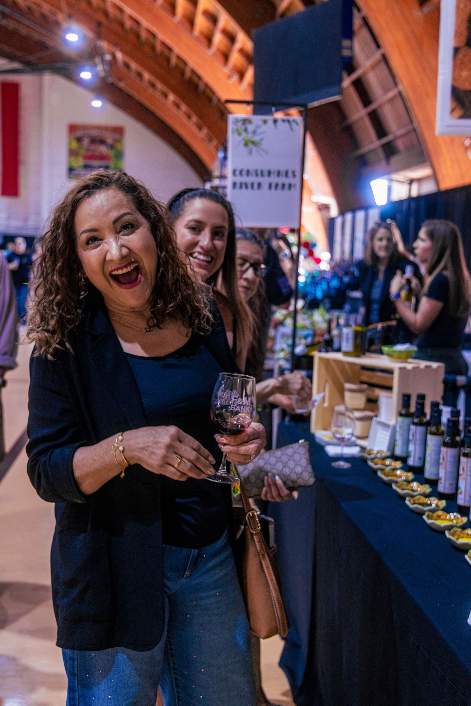 Woman laughing at a wine tasting event, holding a glass of red wine near a vendor table with bottles.
