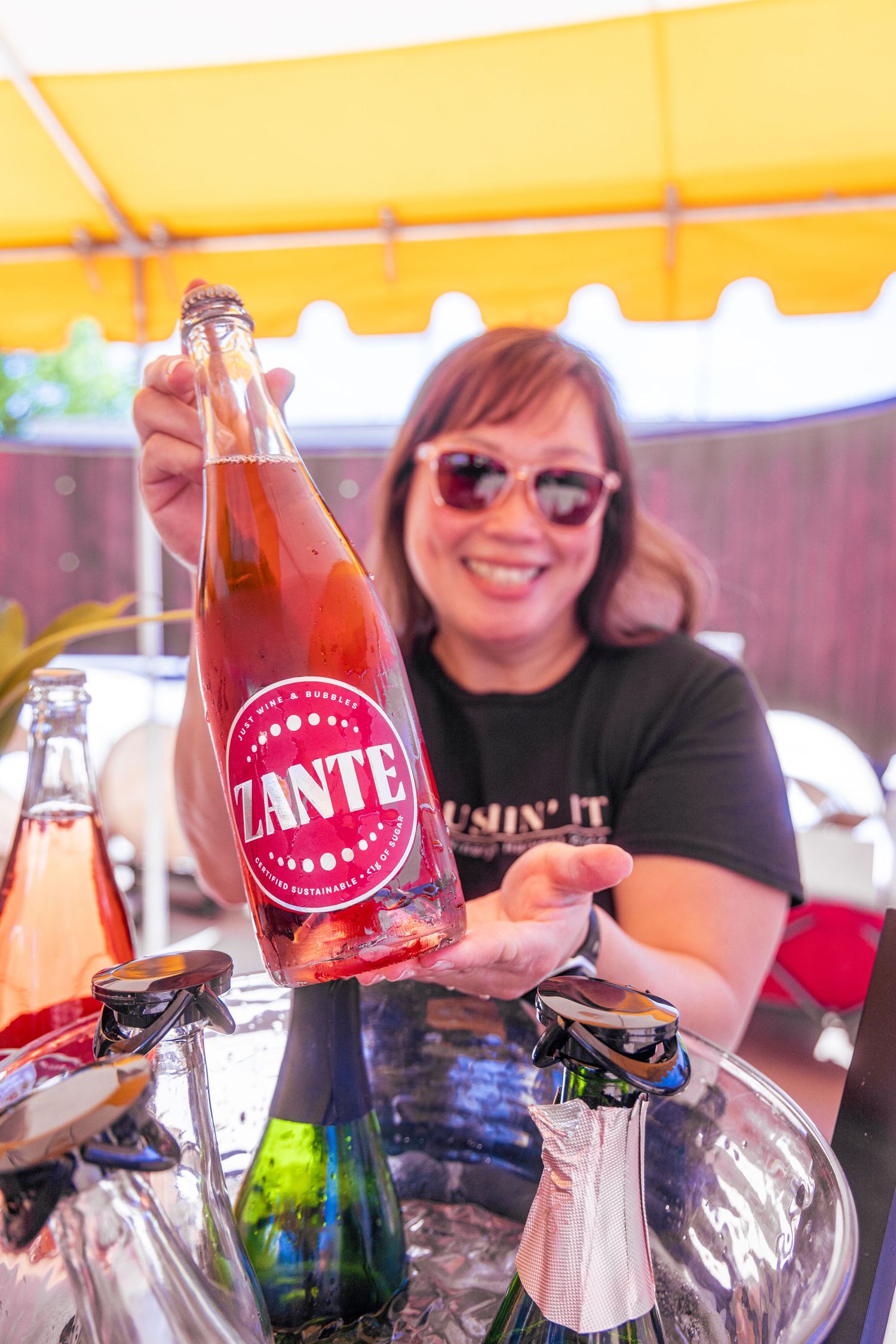 A smiling woman holding a bottle of Zante sparkling rosé wine at an outdoor tasting event.
