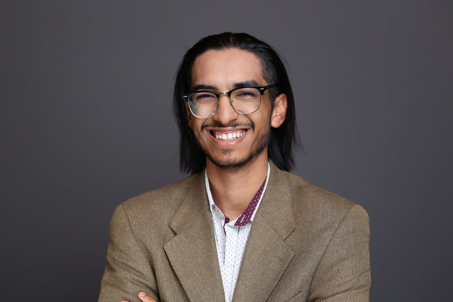 Smiling young man with long hair and glasses in a tan blazer posing with arms crossed.