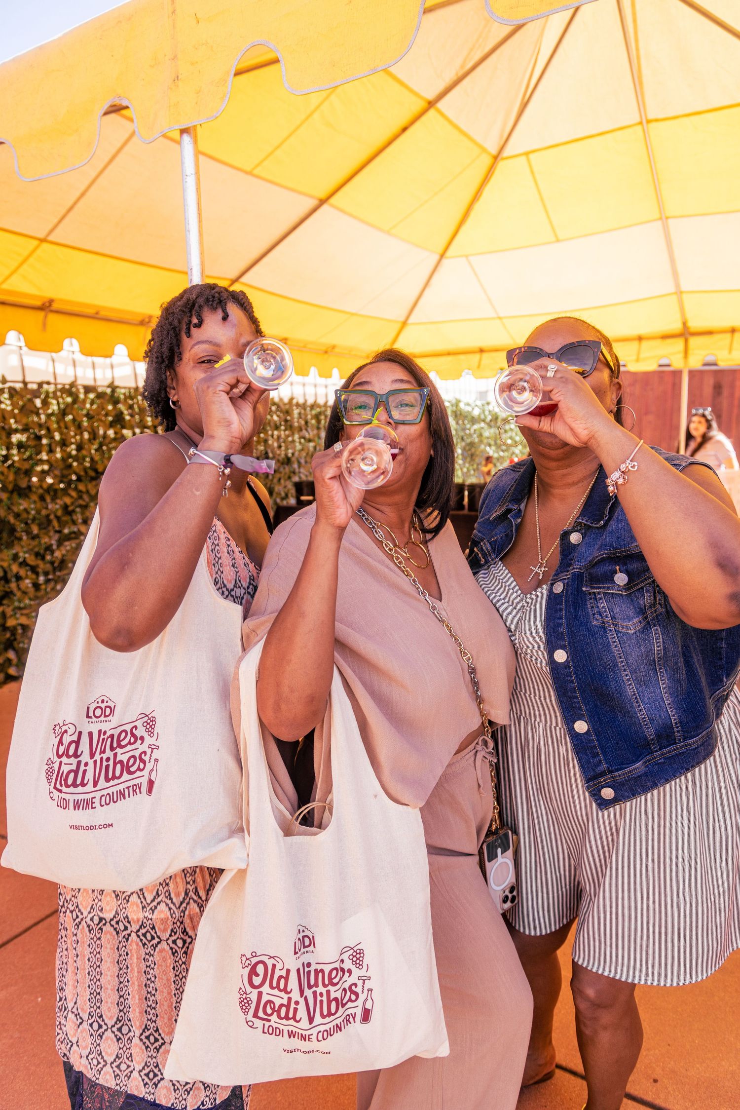 Three women toasting wine at Lodi Wine Country event under yellow umbrella, holding Old Vines Lodi Vibes tote bags.