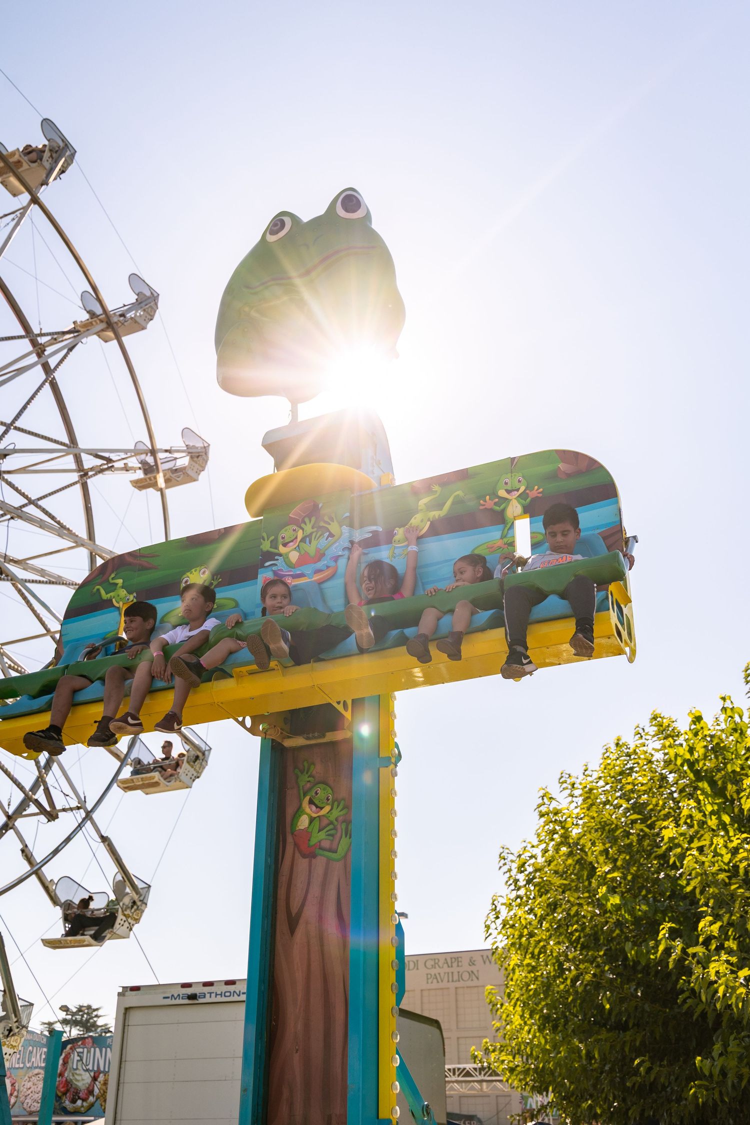 Children ride a colorful frog-themed carnival ride with a Ferris wheel and bright sunburst in the background.
