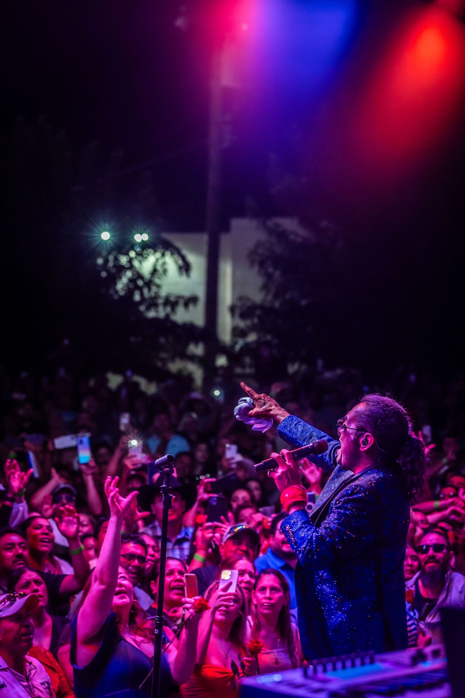 Singer performs on stage with microphone under colorful red and blue lights before a large enthusiastic crowd at night.