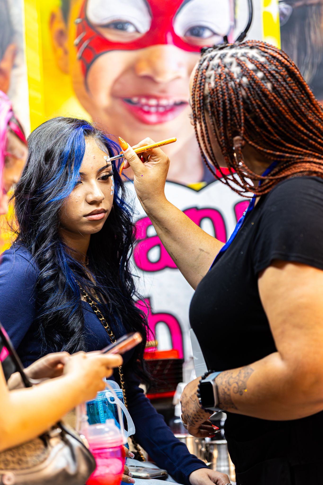 Face painter applying artistic makeup to a young woman with blue hair at a colorful fair booth.