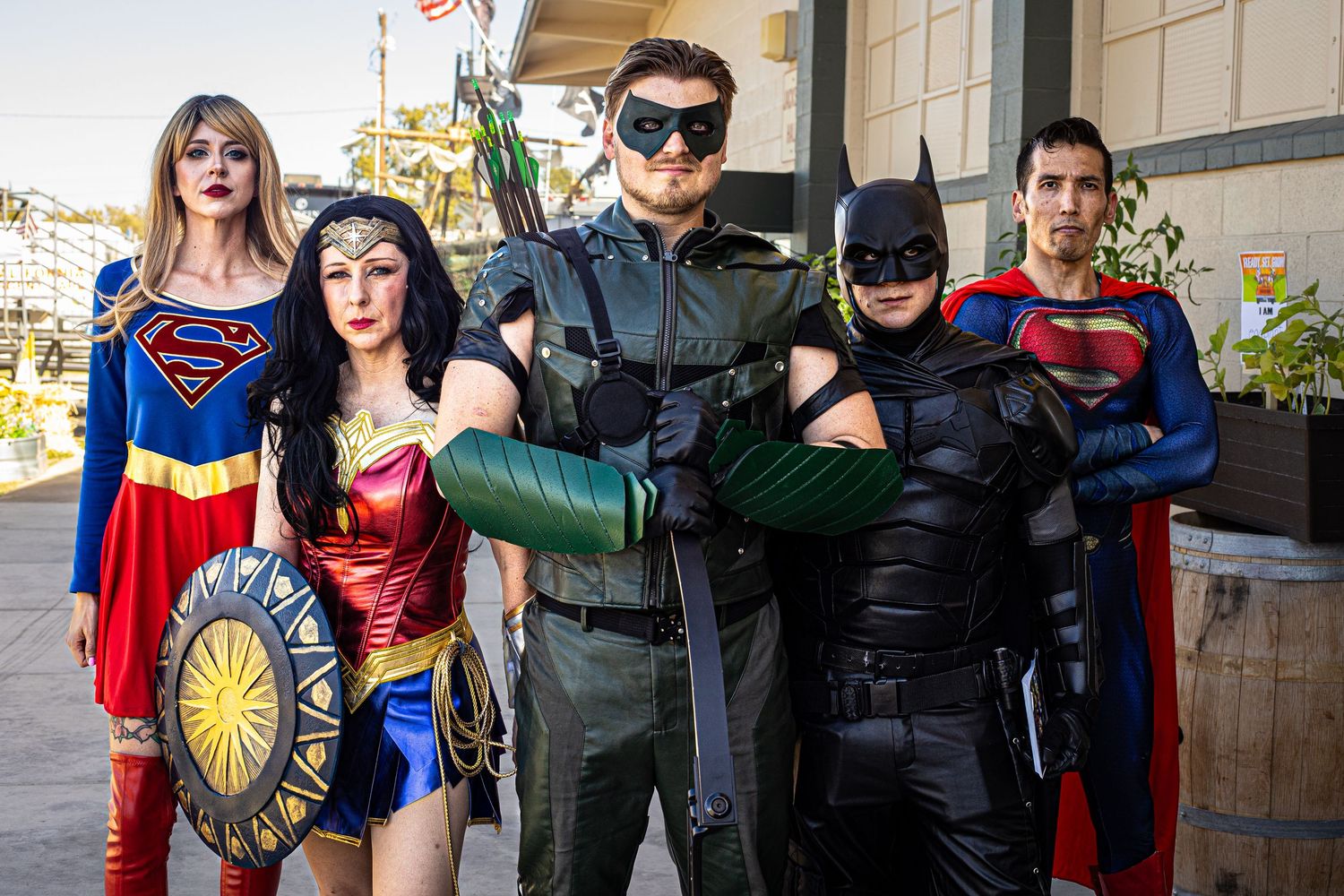Five cosplayers dressed as DC superheroes including Supergirl, Wonder Woman, Green Arrow, Batman, and Superman posing outdoors.