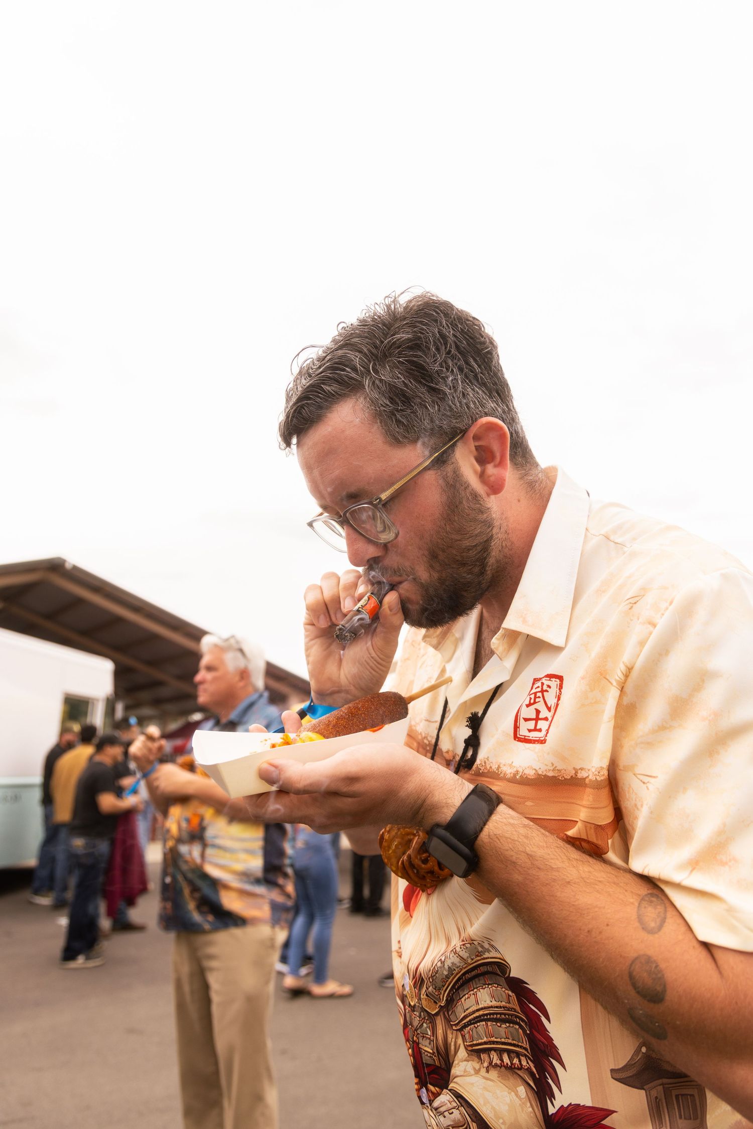 Bearded man in Hawaiian shirt carefully eating food at an outdoor food festival event.